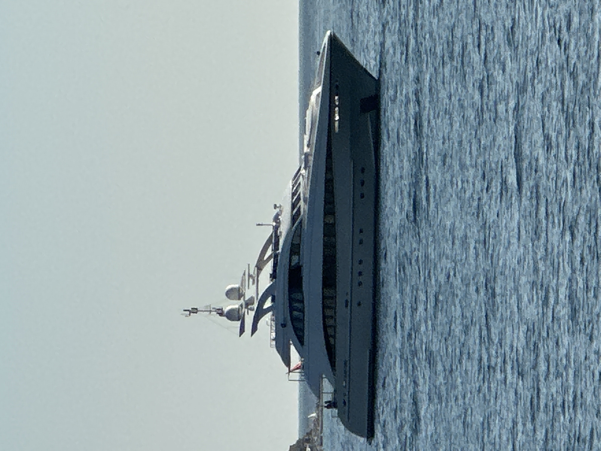 An unidentified, large, white luxury yacht is depicted on a calm, blue sea under a clear sky. The yacht is positioned in the foreground, taking up the majority of the frame, and is seen from its starboard side. It features multiple decks with large windows, sleek lines, and several antennas or communication devices on its upper structure. The yacht appears to be stationary. In the background, a rocky, green landmass, possibly an island or a coastline, is partially visible on the left. The water is textured with gentle ripples. The image is a medium shot, focusing on the yacht and its immediate surroundings. The location is identified as Budva, Montenegro, although there are no specific landmarks to confirm this within the image itself. There are no people or discernible actions occurring on the yacht or in the water.