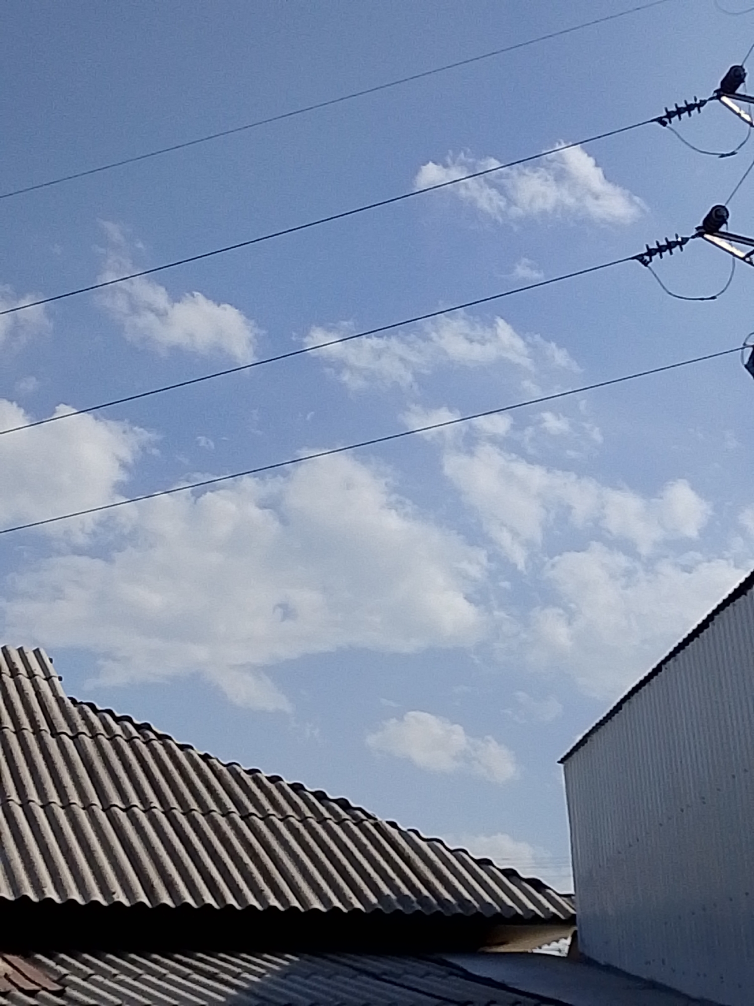 The image shows a close-up of power lines against a bright blue sky with scattered white clouds. In the foreground, the corrugated roofs of two buildings are visible. The roof on the left is made of gray, weathered material and is angled. The building on the right has a white, ribbed metal siding. The scene is an outdoor urban environment, likely during the daytime.