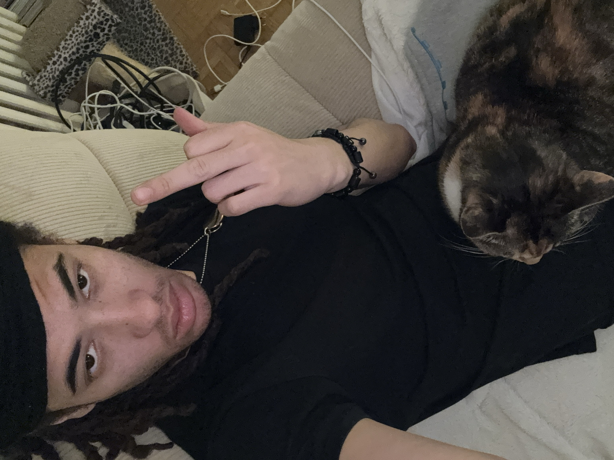 This image depicts an indoor scene set within a domestic environment in Paris, France. A young adult individual with dreadlocks, wearing a black beret or cap, a black short-sleeved shirt, and a silver chain necklace with a light-colored pendant, is seated on a beige ribbed fabric couch or armchair. The individual looks directly into the camera with a serious expression, their right hand raised, displaying an extended middle finger. A black beaded bracelet is visible on their right wrist. Resting on the individual's lap is a tortoiseshell cat, largely visible from its back and right side, facing away from the camera toward the lower right. A white blanket or towel is draped over the individual's lap beneath the cat. In the background, to the right, a white vertical finned radiator is partially visible. Below the radiator, on a light brown wooden floor, there is a cluttered area containing multiple tangled white and black electrical cables, a black power adapter, and two floor coverings, one featuring a leopard print pattern.