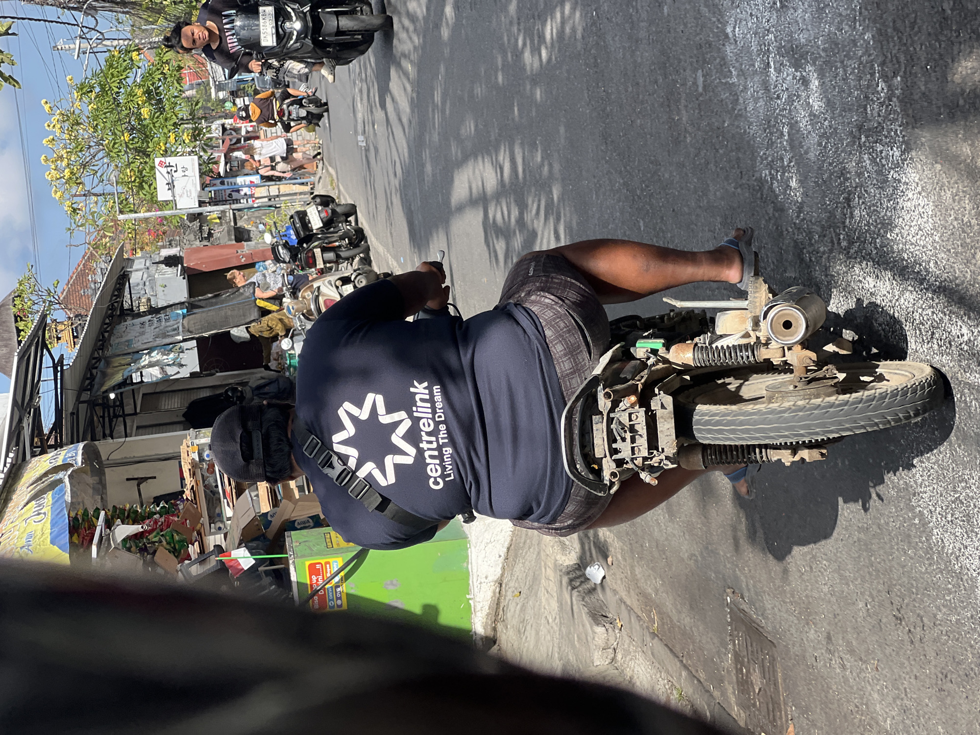 An over-the-shoulder view captures a man riding a motorcycle down a street in Kuta, Indonesia. He is wearing a dark blue t-shirt with a white "centrelink Living The Dream" logo and plaid shorts. The motorcycle is old and appears worn, with visible rust and damage to the rear. The street is paved and appears to be in a commercial area, with shops and stalls lining the sides. Other motorcycles and pedestrians are visible in the background, suggesting a busy urban environment. A small Indonesian flag is planted on a green stand next to a shop on the left side of the street. The sky is clear and sunny, casting shadows on the road.