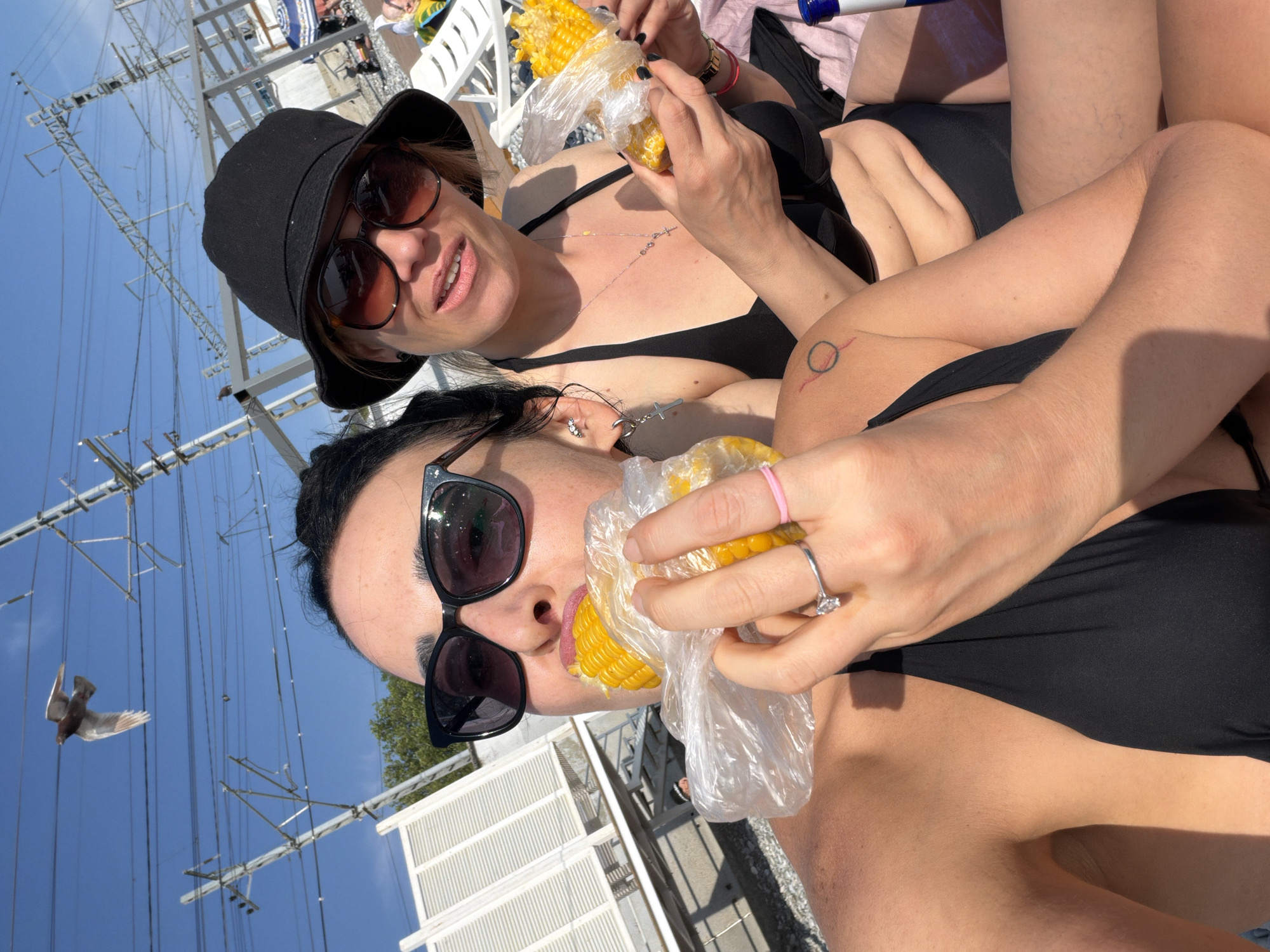 Two women are at a beach in Sochi, Russia, on a sunny day. They are both wearing black bikinis and appear to be enjoying cooked corn on the cob. The woman on the left is closer to the camera and is wearing sunglasses. The woman on the right is wearing a black bucket hat and sunglasses. In the background, there are beach chairs, umbrellas, and power lines overhead. A bird is flying in the sky.