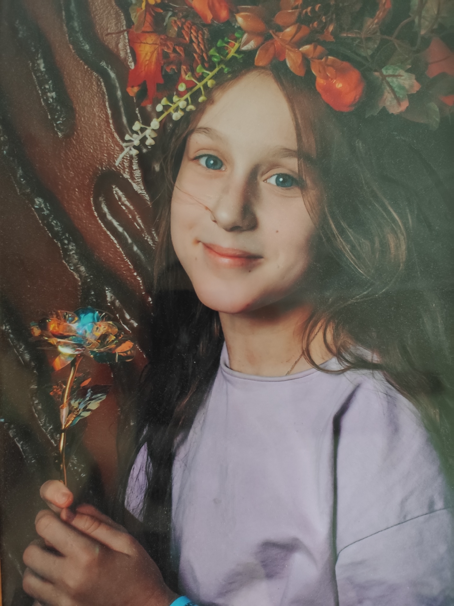 A young girl, likely in her pre-teen or early teenage years, is featured prominently in a portrait-style photograph. She has long, dark brown hair and striking blue eyes, and a gentle smile plays on her lips, conveying a sweet and possibly shy demeanor. She is adorned with an autumn-themed crown or headband, fashioned from red, orange, and brown leaves, interspersed with small white and red berries or flowers.
The girl is wearing a simple, light lavender or pale purple t-shirt. Her hands are positioned in the lower-left portion of the frame, holding a shimmering, iridescent rose-like object. This artificial flower displays a metallic or holographic finish, reflecting a spectrum of colors including blues, oranges, golds, and greens, giving it a magical appearance. A light blue wristband is visible on her left wrist.
The background is a dark brown, featuring an abstract, textured pattern that appears wavy or rippled. The lighting suggests an indoor setting, highlighting the girl and the object she holds. There is no discernible text visible in the image. While no specific landmarks are present, the photograph is set in Moscow, Russia, as per context.
