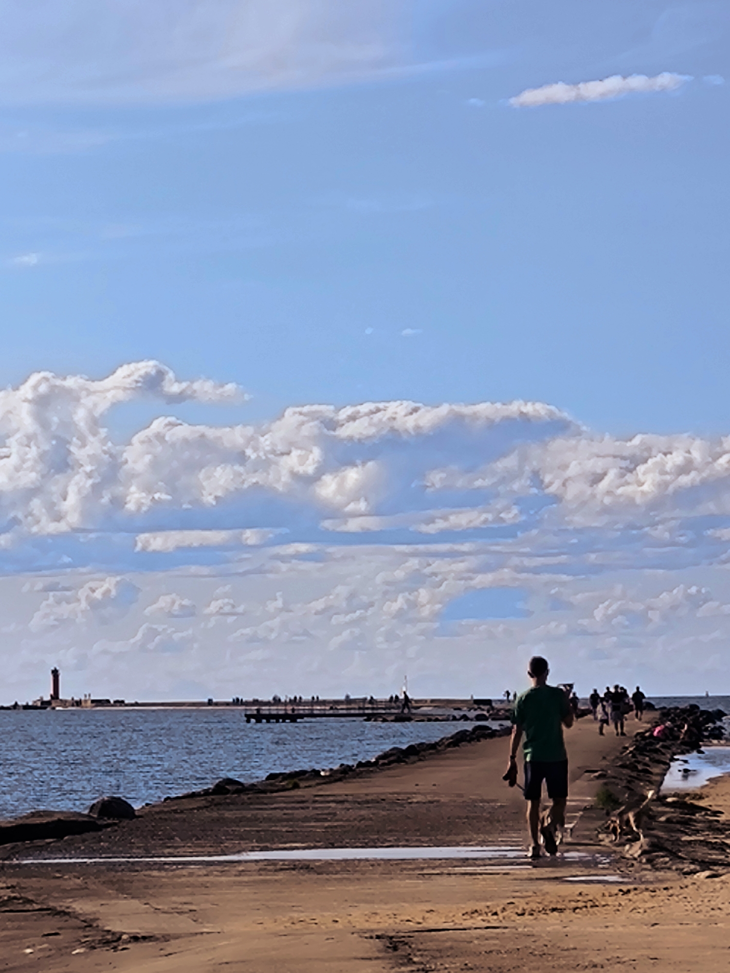 The image depicts a man walking a dog along a concrete pier on a sunny day. The pier extends into a body of water, with a lighthouse visible in the distance. Several other people are also present on the pier, some walking and others standing. The sky is blue with scattered white clouds. This scene appears to be located at the Daugava River estuary in Riga, Latvia, given the presence of a lighthouse and pier characteristic of the area.