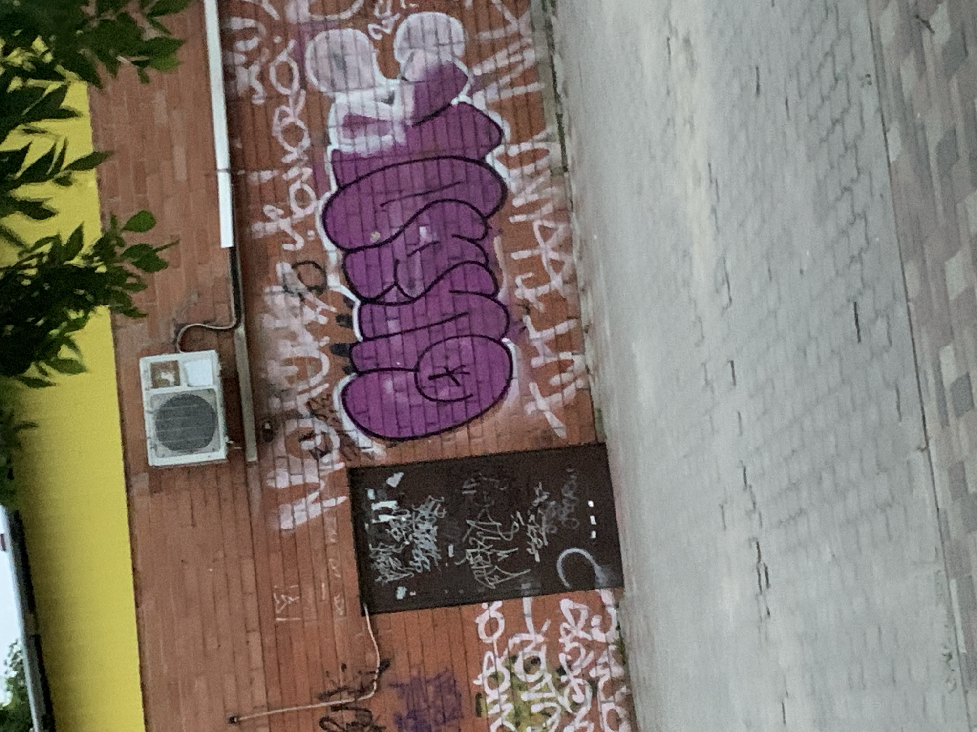 A street scene in Stavropol, Russia, shows a brick building wall covered in graffiti, with a distinct purple graffiti tag dominating the view. An air conditioning unit is mounted on the wall near the graffiti. A black rectangular object with additional graffiti is visible below the purple tag. The building is situated next to a paved sidewalk. A tree is visible on the left.