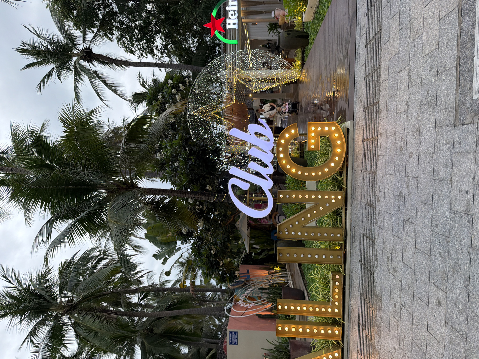 The image depicts a nighttime scene outdoors, likely in Nha Trang, Vietnam, with a focus on a brightly lit entrance to a venue. Large, illuminated marquee letters spell out "CLUB" in white, positioned in the foreground and center. To the left, other illuminated marquee letters, in a golden hue, are partially visible, spelling out "ALLING". Behind these, a neon arch structure with lights in a circular pattern is visible, leading towards the entrance of a building.
The building itself appears to be a restaurant or bar, with white canopies and outdoor seating. A prominent Heineken logo, featuring a red star and green branding, is visible on the right side of the building's facade. The ground in front of the entrance is a wooden boardwalk, which appears wet, suggesting recent rain. This boardwalk is lined with lush green plants and potted trees.
In the background, numerous palm trees are visible, characteristic of a tropical or subtropical coastal location like Nha Trang. The sky is overcast and dark, indicating it is evening or night. A person is seen pushing a stroller with a child on the wet boardwalk, walking towards the illuminated entrance. The overall impression is of a lively, festive atmosphere at a beachside establishment.
