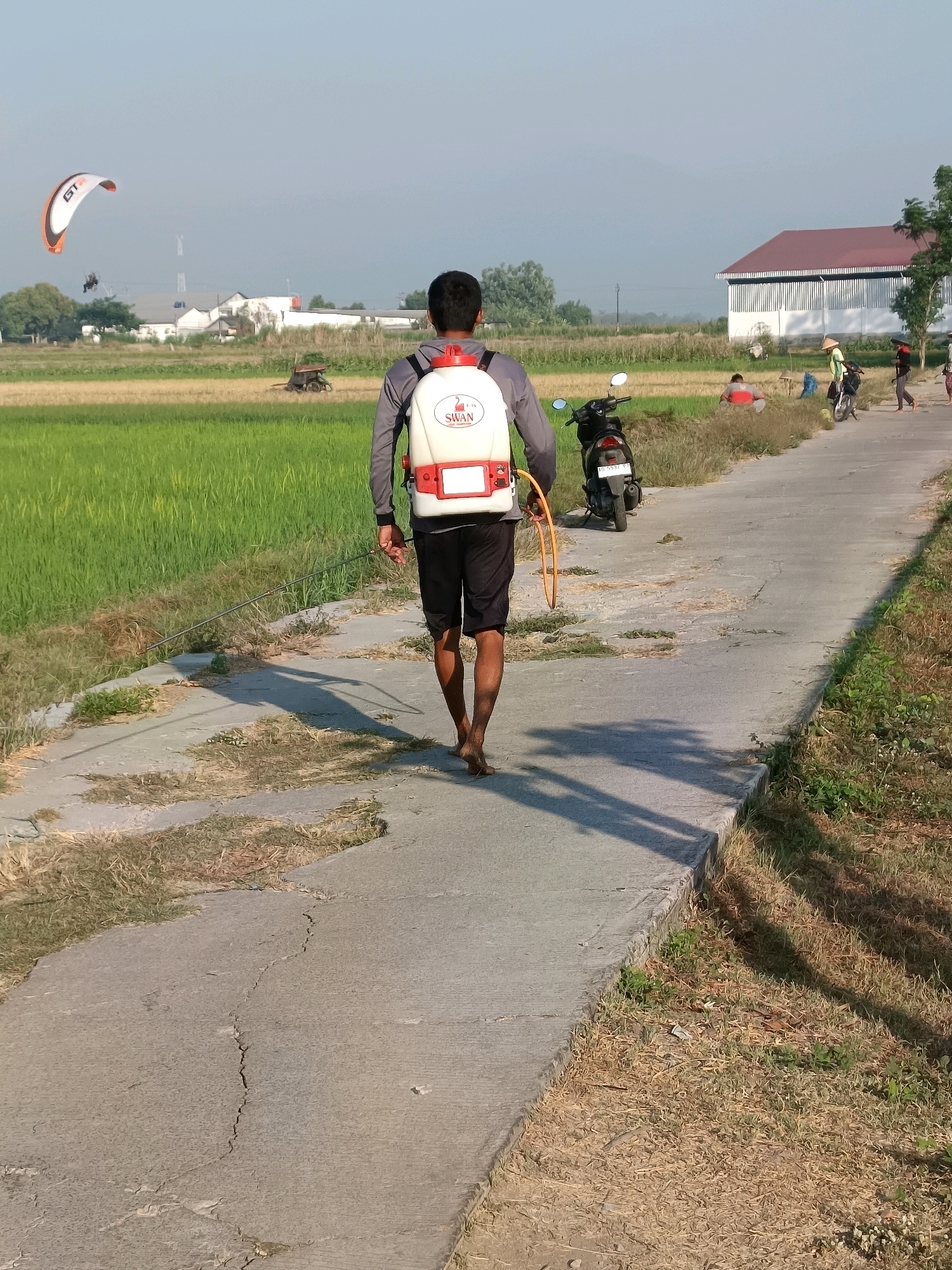 A man walks away from the camera on a paved path in Geneng, Indonesia, carrying a white and red sprayer backpack with the "SWAN" logo. He is wearing a gray long-sleeved shirt and black shorts, and is barefoot. To his left is a lush green rice paddy. To his right, a black motorcycle is parked on the side of the path. In the distance, several people are visible in and around a building with a red roof. A paraglider is in the sky above the rice paddy. The sun is casting long shadows across the path.