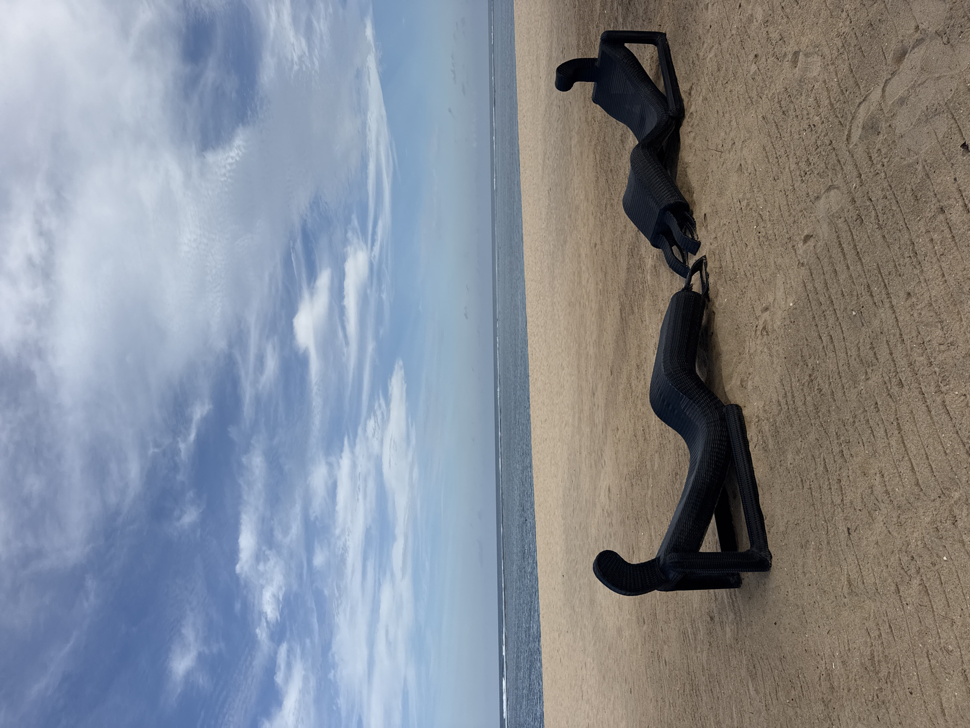 A wide shot shows two black, abstract, human-shaped lounge chairs on a sandy beach. The chairs are positioned facing each other, connected by a flexible black material. The beach is a light brown color with visible sand patterns from previous footsteps or grooming. The ocean is visible in the background, a deep blue, with a thin white line of waves breaking near the shore. The sky above is a clear blue with scattered, wispy white clouds. The scene is set in Kuta Selatan, Indonesia, a popular tourist destination known for its beaches.