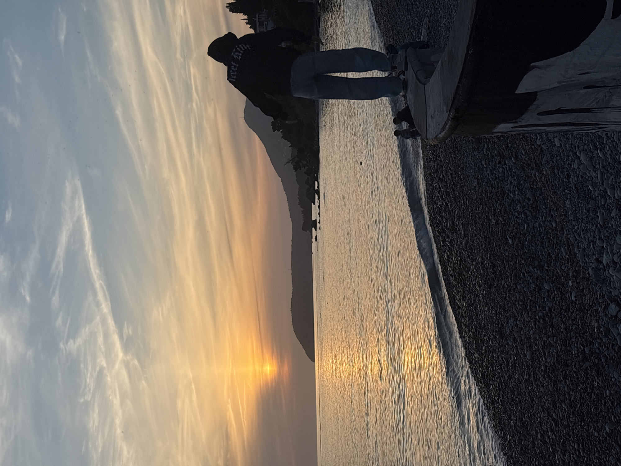 A horizontal image shows a person standing on a beach at sunset. The person is wearing a black hoodie and blue jeans, facing away from the camera and looking at the sunset. The beach has a rocky surface. The water reflects the light of the sunset, creating a shimmering effect. In the background, a mountain range is visible. The sky is a mix of blue, orange, and yellow hues, with wispy clouds scattered across the expanse. This scene is likely located in the city of Bar, Montenegro, given its coastal location and mountainous backdrop.