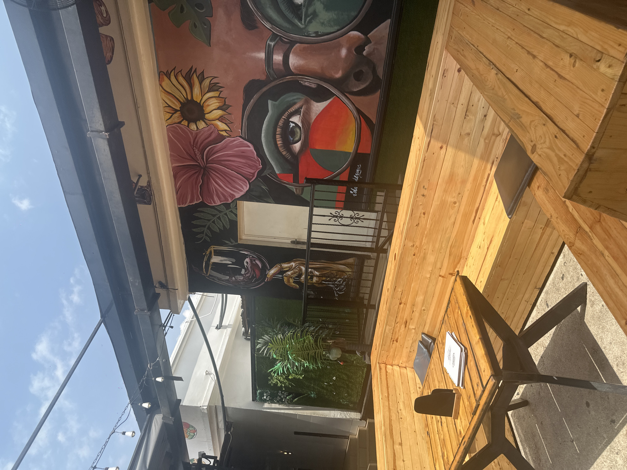 This is an exterior shot of a patio dining area with a vibrant mural on the back wall. The mural features a stylized depiction of a woman's face with large glasses, adorned with flowers and abstract shapes. To the left of the mural, a green wall with artificial plants and a palm tree is visible, with a depiction of a hand holding a wine glass above it. In the foreground, a wooden picnic table with a cross-shaped base is set on a concrete floor. A menu and a black sign holder sit on the table. A large, L-shaped wooden bench seating area surrounds the table. String lights hang from a black awning overhead. The scene suggests a restaurant or bar setting, possibly in the city of Howayi, Nigeria, given the context. The sky above is blue with scattered clouds.