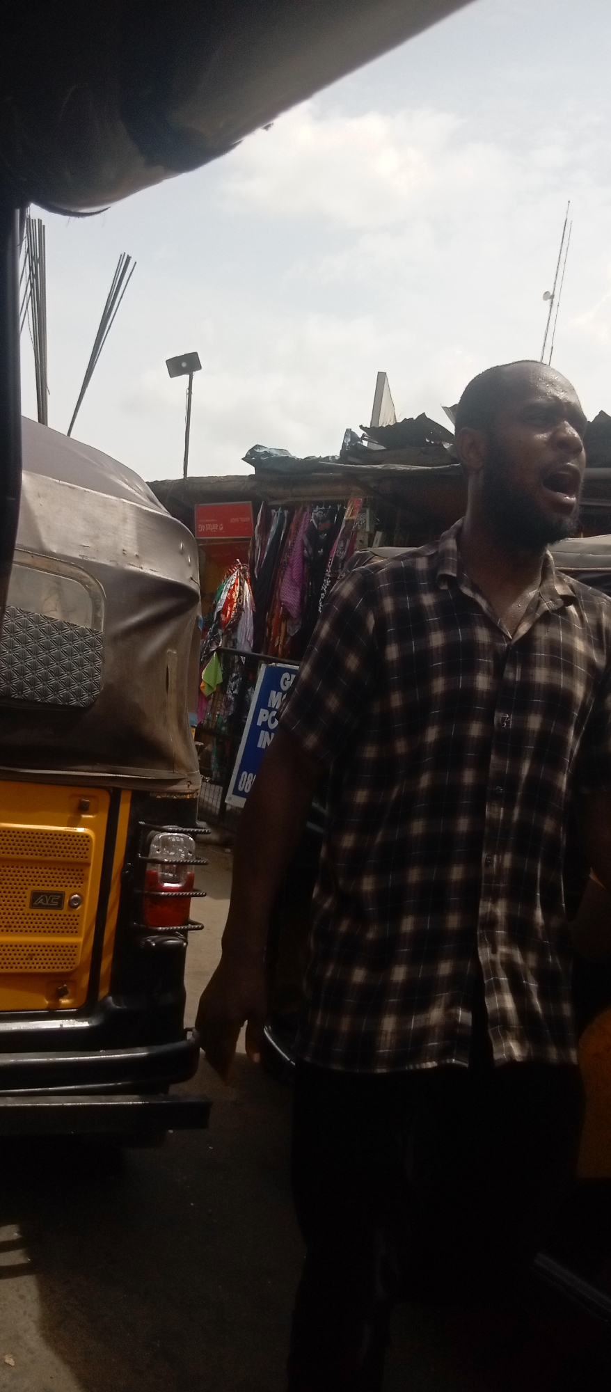 The image captures a moment in Onitsha, Nigeria. A man with dark skin and a beard stands outdoors, wearing a checkered short-sleeved shirt and dark pants. His mouth is open as if he is speaking or shouting. The man appears to be sweating. Beside him is a yellow and black tricycle vehicle (keke). In the background, there is a shop selling clothing. The sky is overcast. The photo is taken at street level.