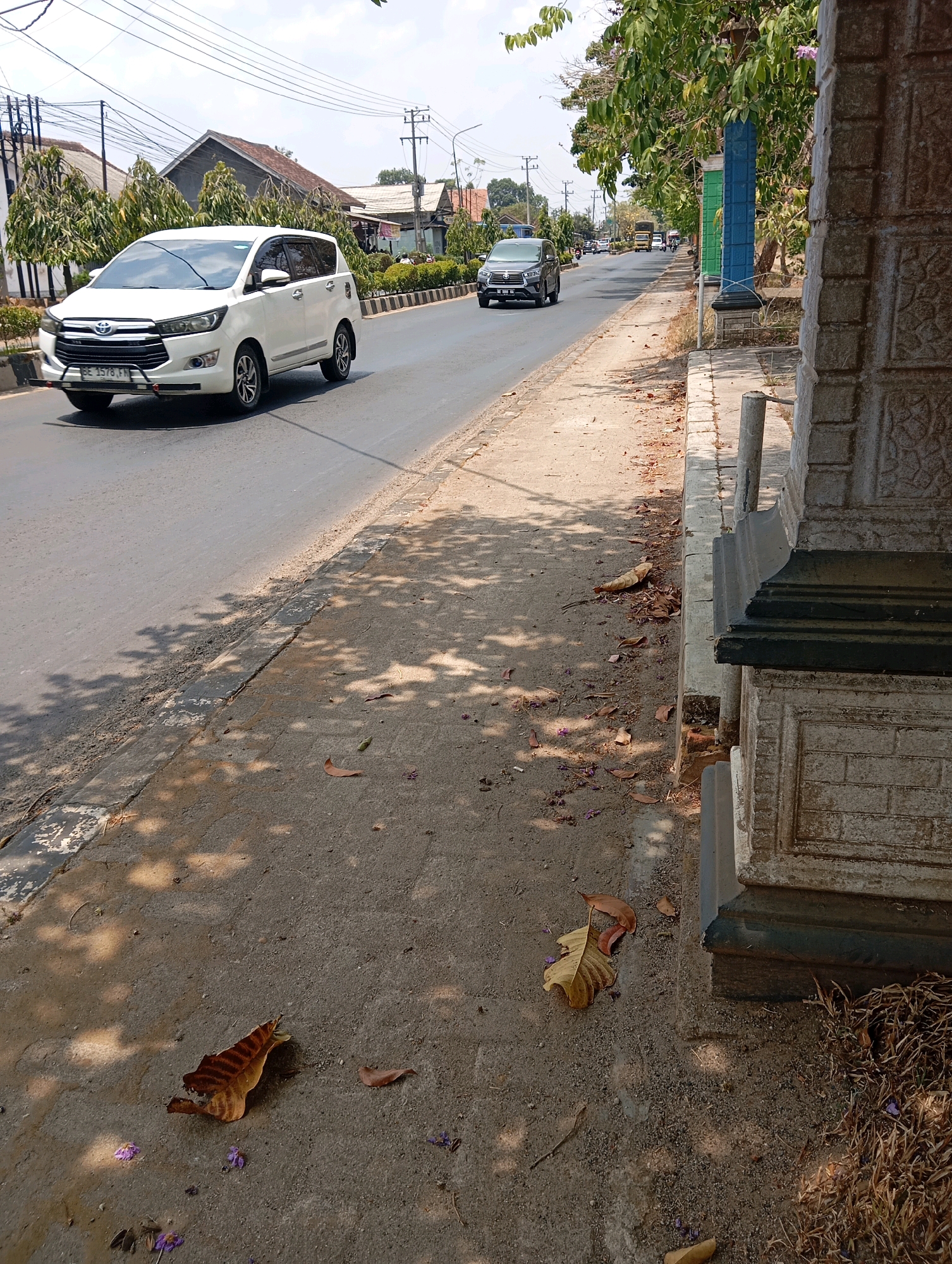 This image depicts a street scene in Metro, Indonesia, with a focus on the sidewalk and the adjacent road. A white Toyota Innova minivan is driving on the road, followed by a dark-colored SUV. The sidewalk is covered in fallen leaves and some small purple flowers. Trees line the sidewalk, casting dappled shadows on the ground. In the foreground, a portion of a stone pillar with intricate carvings is visible, suggesting it might be part of a building or a decorative structure. The overall environment suggests a sunny day in a semi-urban or suburban area, with evidence of vegetation and residential buildings in the background.