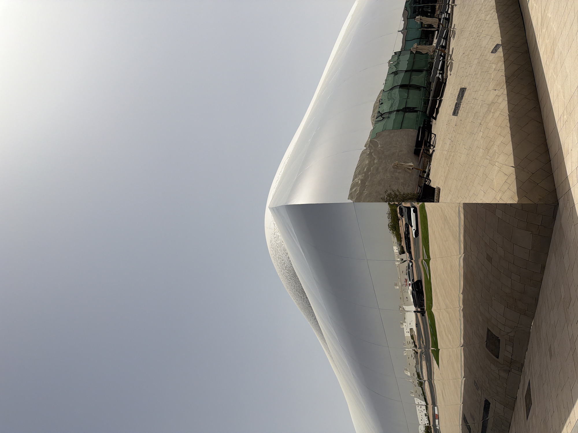 A low-angle, almost vertically oriented photograph reveals a section of the National Museum of Qatar, situated in Al Rayyan. The building's exterior is predominantly white, with a polished finish that reflects the adjacent urban landscape. The reflection includes buildings, vehicles, and the ground level surfaces of the cityscape. The sky above is pale gray, suggesting an overcast or bright, diffused lighting. The structure's architecture is characterized by its curved, intersecting planes. The building abuts a wall covered in beige stone blocks, and the image is taken from a low angle, focusing on the building’s unique structure and its reflective qualities. There are no people visible in the image itself, but the reflections within the building’s surface suggest the presence of pedestrians and vehicular traffic in the surrounding area.