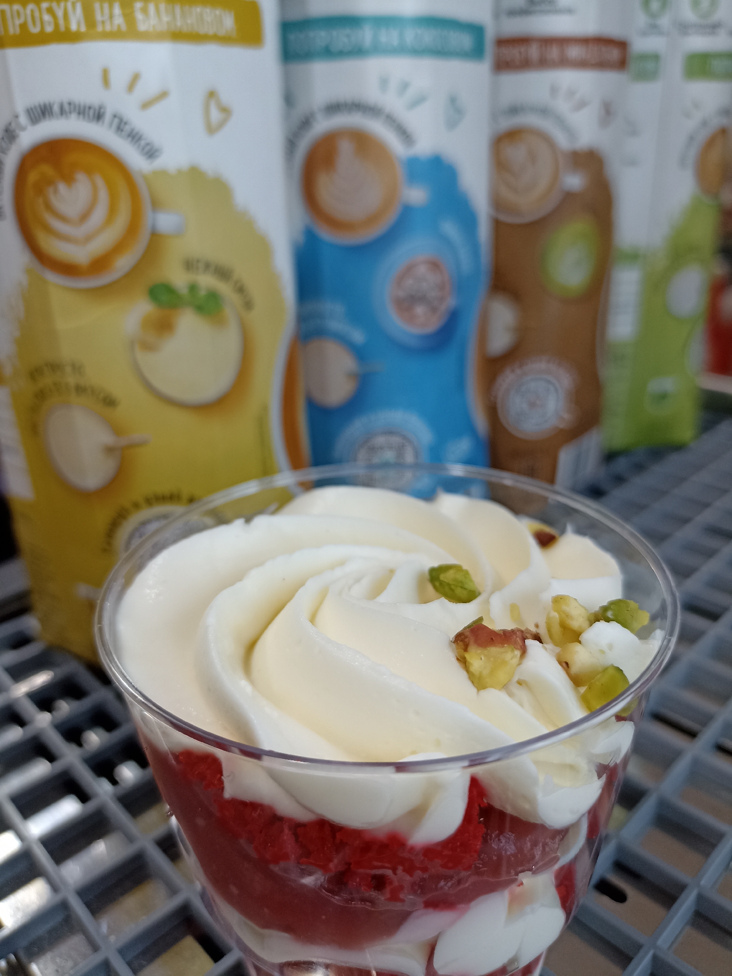 A close-up shot displays a dessert in a clear plastic cup with a swirled topping of white frosting adorned with chopped pistachios. Below the frosting, layers of red, possibly strawberry or raspberry-flavored filling, and additional white filling are visible. The dessert sits on a gray metal grating. Behind the cup, out of focus, are four containers, possibly food or beverage products. The container on the left is yellow with images of a latte and a pudding-like dessert. The other three containers are blue, brown, and green, each with a latte image. The location is marked Unknown, Unknown. The setting and contents suggest a commercial environment such as a bakery or cafe.