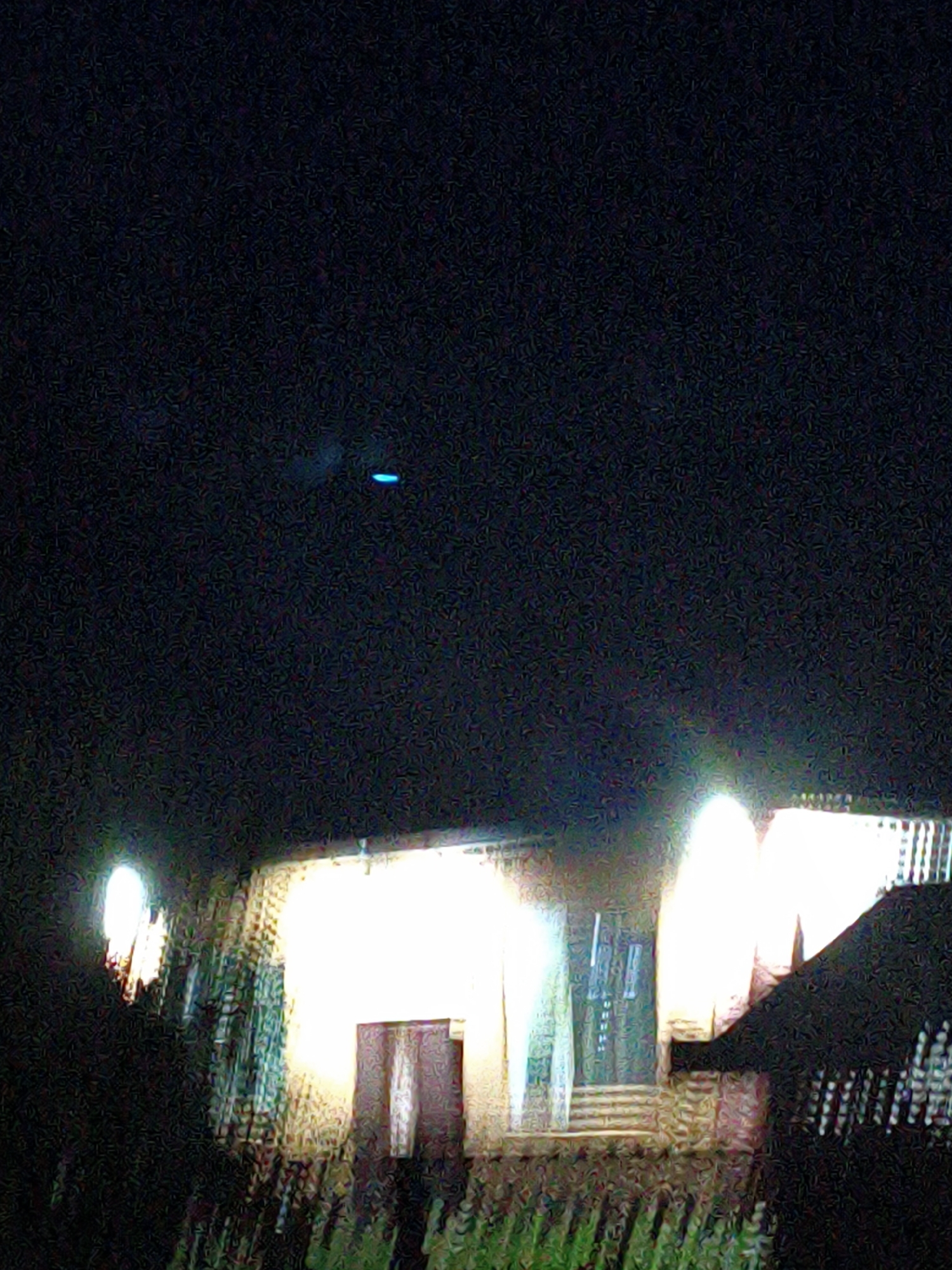 The image depicts a dark, outdoor scene at night, likely in Ustyvytsia, Ukraine. In the upper portion of the image, a faint, horizontal blue light is visible against the black sky. The lower portion of the image shows a brightly lit structure, possibly a building or house. The light emanates from windows or illuminated panels, casting a warm glow. In the foreground, there are shadowy, indistinct shapes that could be trees or vegetation. The overall image quality is grainy, suggesting a low-light photograph taken with a low-resolution camera or under challenging lighting conditions. There are no visible people or specific objects that can be clearly identified. The scene appears to be a residential or public area illuminated at night.