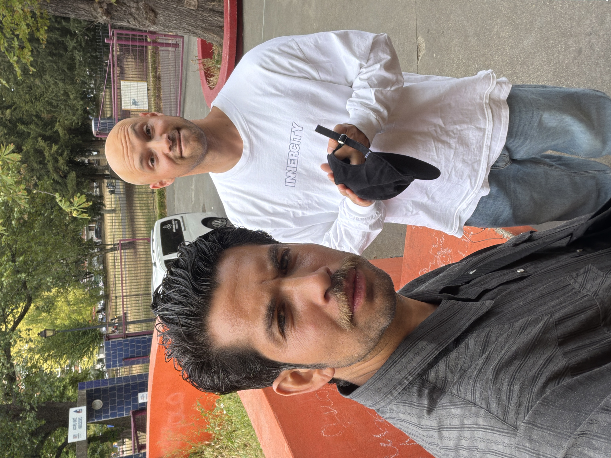 Two men are pictured in an outdoor setting. The man in the foreground, with dark, slicked-back hair and a mustache, is wearing a dark, pinstriped button-down shirt. He is looking directly at the camera with a neutral expression. The man behind him, who is bald and has stubble, is wearing a white long-sleeved shirt with the word "INERCITY" printed vertically on the chest in an outline font. He is holding a black baseball cap in his right hand. The background suggests an urban environment with trees, a fence, and a building with blue tiled accents. There is a red concrete structure in the foreground, possibly a bench or planter. The lighting indicates it is daytime, with natural light illuminating the scene. The overall mood appears casual and relaxed. No specific event is clearly depicted, but it seems to be a candid moment captured outdoors.