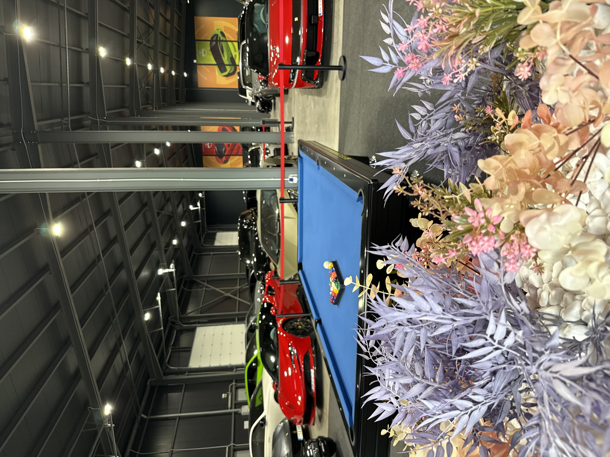 This media file depicts a spacious indoor setting, likely a luxury car showroom, a private collection garage, or an event space, characterized by an industrial aesthetic combined with elements of leisure and display.
In the immediate foreground on the right, a large arrangement of artificial flowers and foliage is prominently featured. The arrangement includes light purple (lavender) leafy branches, various shades of pink flowers, and cream or light peach blossoms, some of which are blurred due to their closeness to the camera.
The midground showcases a blue-felt pool table with a rack of billiard balls. To the left of the pool table, a collection of high-end sports cars is displayed. A vibrant red sports car is most conspicuous, followed by black, white, and a distinct bright green car in the distance. These vehicles appear to be part of an organized exhibit, with black stanchions and red ropes partially visible, suggesting a roped-off area.
The background reveals a high ceiling constructed from exposed metal beams and corrugated panels, indicative of a large, industrial-style building. Multiple bright, artificial lights illuminate the entire space. On the far wall, several large, glowing display panels are visible, one featuring a graphic of a green car or car component against an orange background, and another with a red graphic.
No people are visible in the image, and no specific activity is taking place; the scene appears set up for viewing or future events. The lighting is entirely artificial, suggesting an indoor environment regardless of the time of day. No readable text is clearly discernible on any of the cars, displays, or other objects.