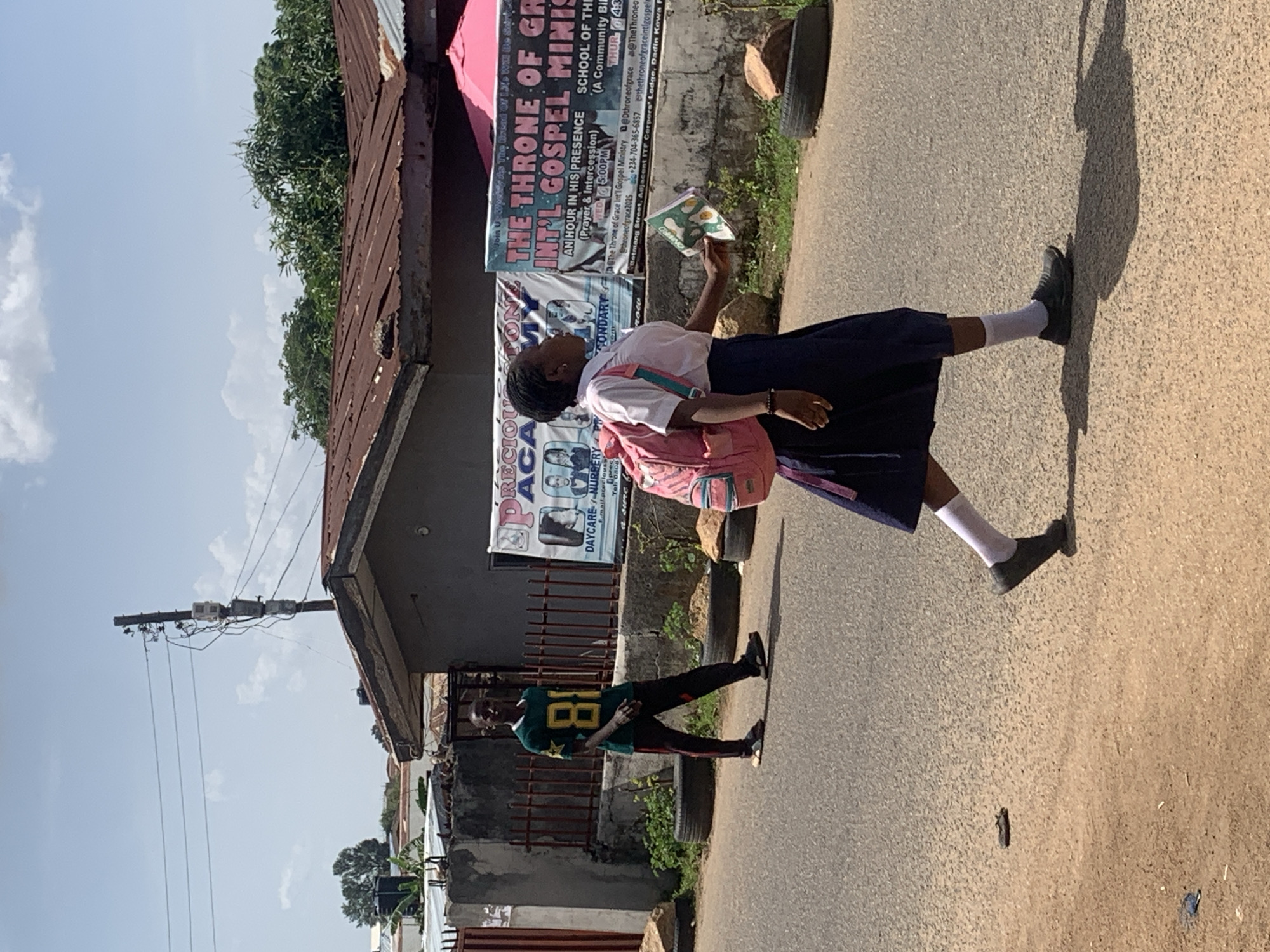The image depicts an outdoor street scene in Jos, Nigeria, captured during bright daylight hours, evidenced by the clear sky and distinct shadows. The setting features a paved road bordered by buildings with weathered corrugated metal roofs and some green foliage.
In the foreground, a young person, likely a student, walks along the asphalt road. They are dressed in a school uniform comprising a white shirt, a navy blue skirt, white socks, and dark shoes, and carry a pink backpack. This individual is holding a small book or pamphlet, possibly reading or examining its contents as they proceed. Their shadow is prominently cast on the road.
Further back on the street, another person is visible, wearing a green jersey with the large yellow number "81" on the front, and dark trousers. This individual appears to be standing near a low wall or fence. A power pole with overhead wires is also visible against the bright, partially cloudy sky.
Banners and posters adorn the walls of the buildings. Visible text includes "THE THRONE OF GRACE INT'L GOSPEL MINISTRY," advertising "AN HOUR IN HIS PRESENCE (Prayer & Intercession)" with timings "WED 5.00PM" and "THUR 4:30PM." A phone number, "+234-704-365-6857," and social media handles like "@Dthroneofgrace" are also present. Other signs mention "DAYCARE NURSERY" and "SCHOOL OF THE..." and reference "Betmang Street, Adjacent ITF Corpors' Lodge, Dadin Kowa P," confirming the local context. The book held by the student shows the partial text "ONFRO". The overall scene suggests a glimpse into daily life within a bustling urban or semi-urban environment.