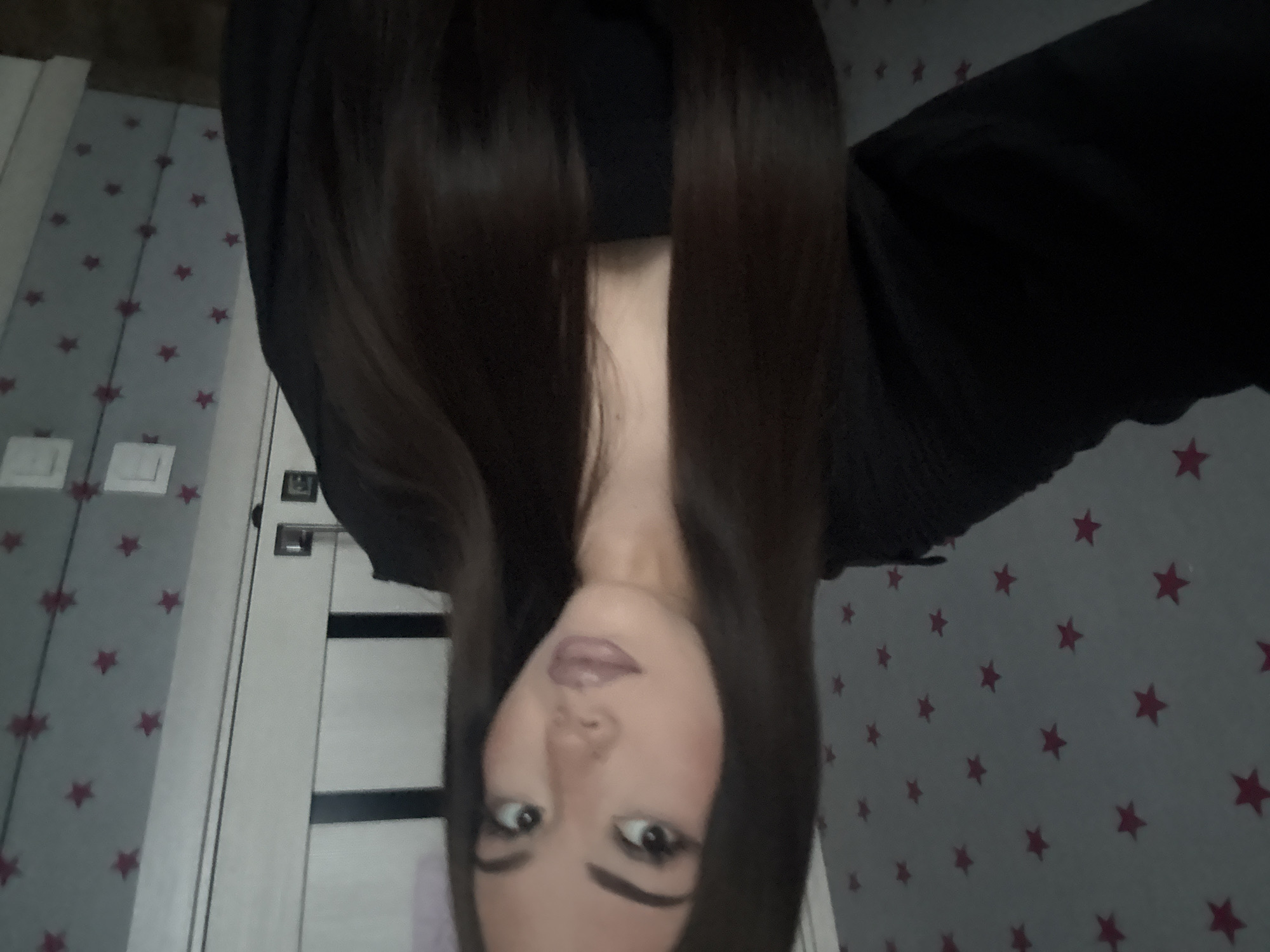 A young woman with long, dark brown hair is pictured indoors, appearing to be in a bedroom or hallway. The background features a gray wall with a repeating pattern of small, dark pink stars. To the right of the woman, a white door with dark horizontal inlays is visible, along with light switches and what appears to be a reflection of the room in a mirror or glossy surface. The woman is wearing a black, loose-fitting top. Her facial features are visible; she has on makeup, including defined eyebrows, eyeliner, and lip gloss. The lighting is dim, and the overall image quality is slightly grainy. The location is identified as the city of Naberezhnye Chelny, Russia.