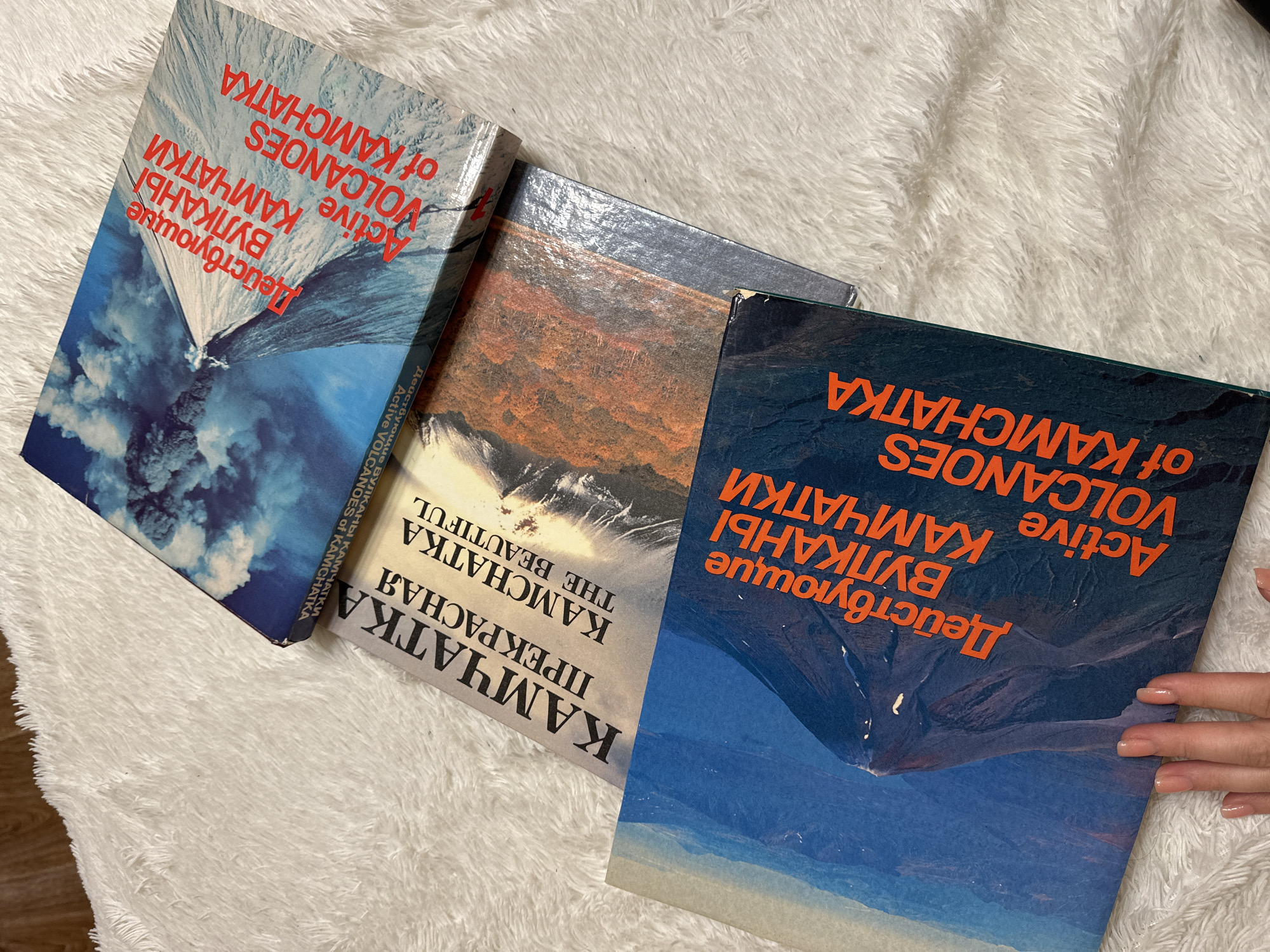 The image shows three books titled "Active Volcanoes of Kamchatka" and "Kamchatka the Beautiful" lying on a white, fluffy surface. The books are stacked slightly on top of each other. The book in the foreground is partially held by a hand with manicured nails. The text on the books is in both English and Russian. The book covers feature landscape images, presumably of volcanoes.