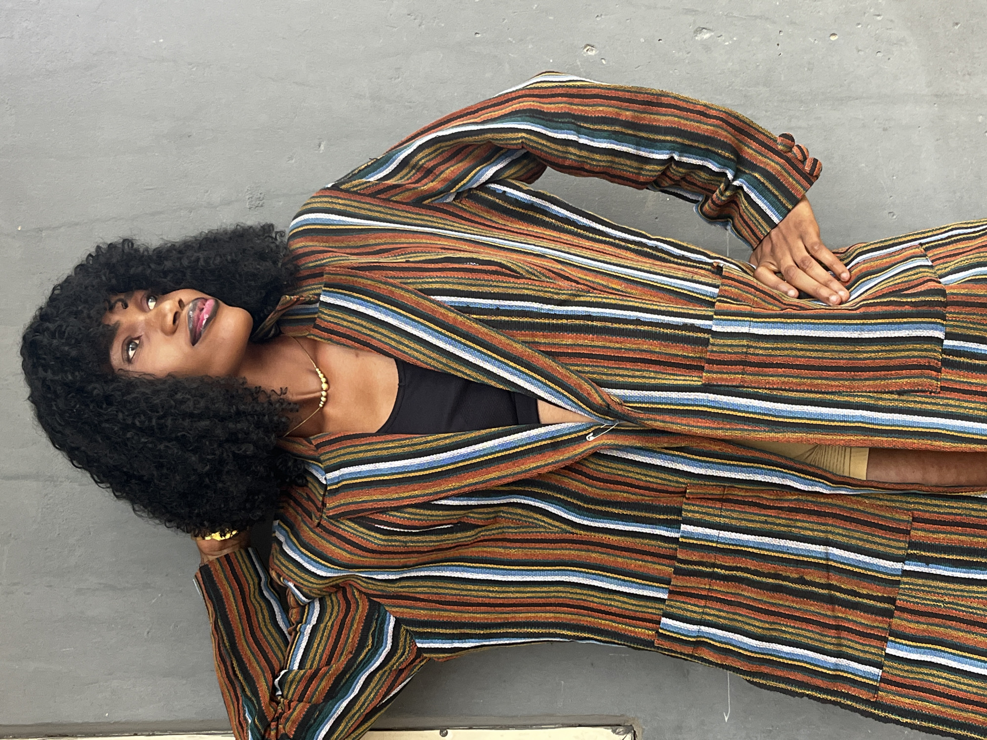 A woman with dark curly hair is lying on a gray concrete surface, looking upwards. She is wearing a striped suit, unbuttoned to reveal a black tank top underneath, along with beige shorts. Her left hand rests on her hip, and a gold bracelet is visible on her left wrist. The scene's location, Badagry, Nigeria, is not evident from the image alone. The image appears to be a fashion or portrait shot.