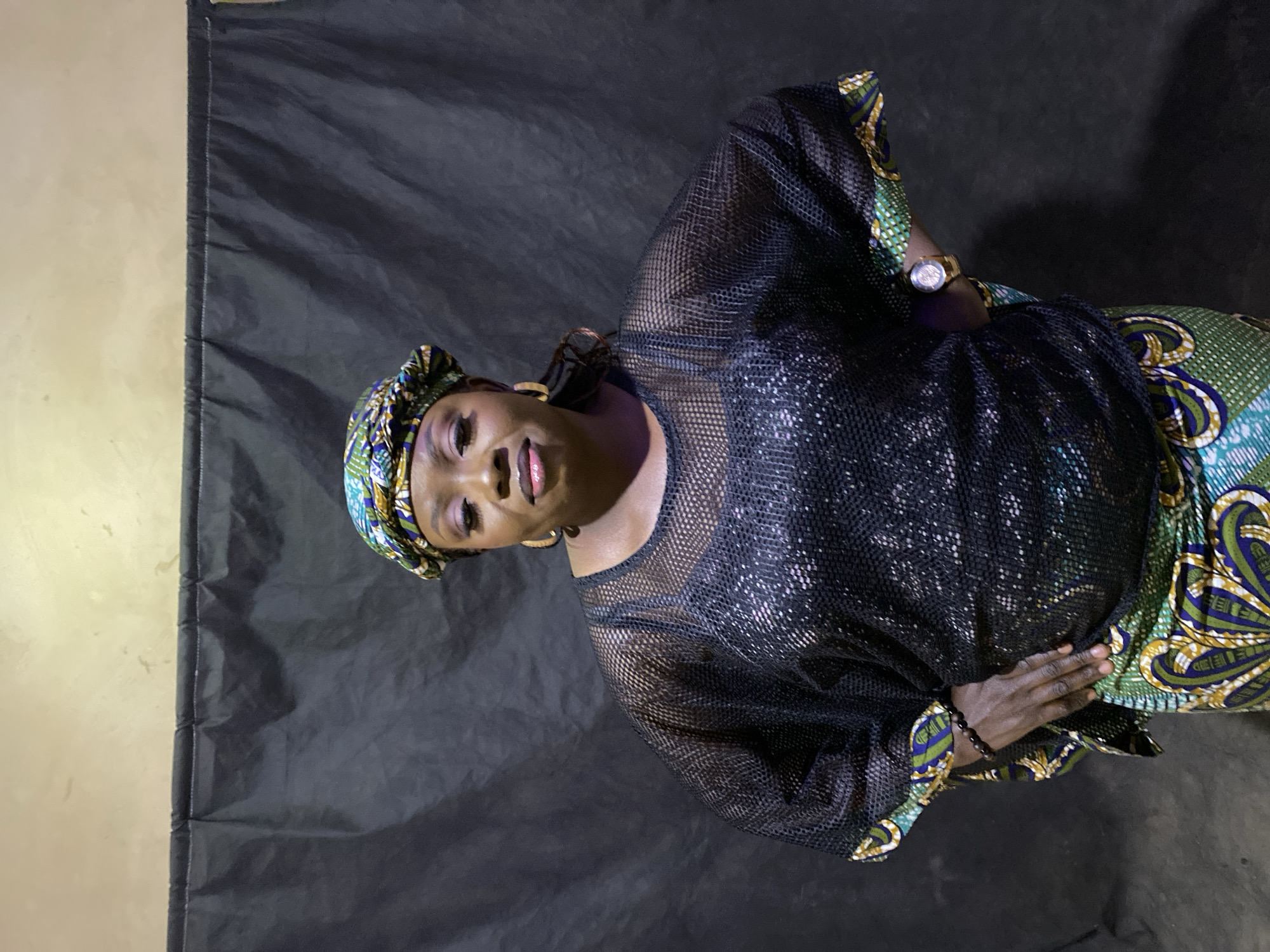 The image displays a head-and-torso portrait of a woman, oriented ninety degrees counter-clockwise. She has dark skin and is looking directly at the camera with a slight, gentle smile. Her face is made up, featuring defined brows, eyeliner, and pinkish lipstick.
She wears a colorful headwrap (gele) with patterns in shades of green, blue, yellow, and black. Her attire includes a black mesh top, which appears to have sequins or a shimmery layer underneath, and a vibrant patterned fabric garment, likely a wrapper or skirt, visible from her waist down, featuring similar green, blue, and yellow designs. She accessorizes with large gold-toned hoop earrings, a metallic wristwatch on her left wrist, and a dark beaded bracelet on her right wrist. Her right hand is subtly placed on her patterned garment.
The woman is posed against a dark, possibly black or dark grey, fabric backdrop, which shows some folds and creases. A light-colored wall is visible on the far left side of the frame, suggesting the backdrop does not cover the entire background. The lighting is focused, illuminating the subject. No specific activity beyond posing for the photograph is depicted. The indoor setting and traditional attire are consistent with the location context of Kuru Dungus, Nigeria. There is no visible text.