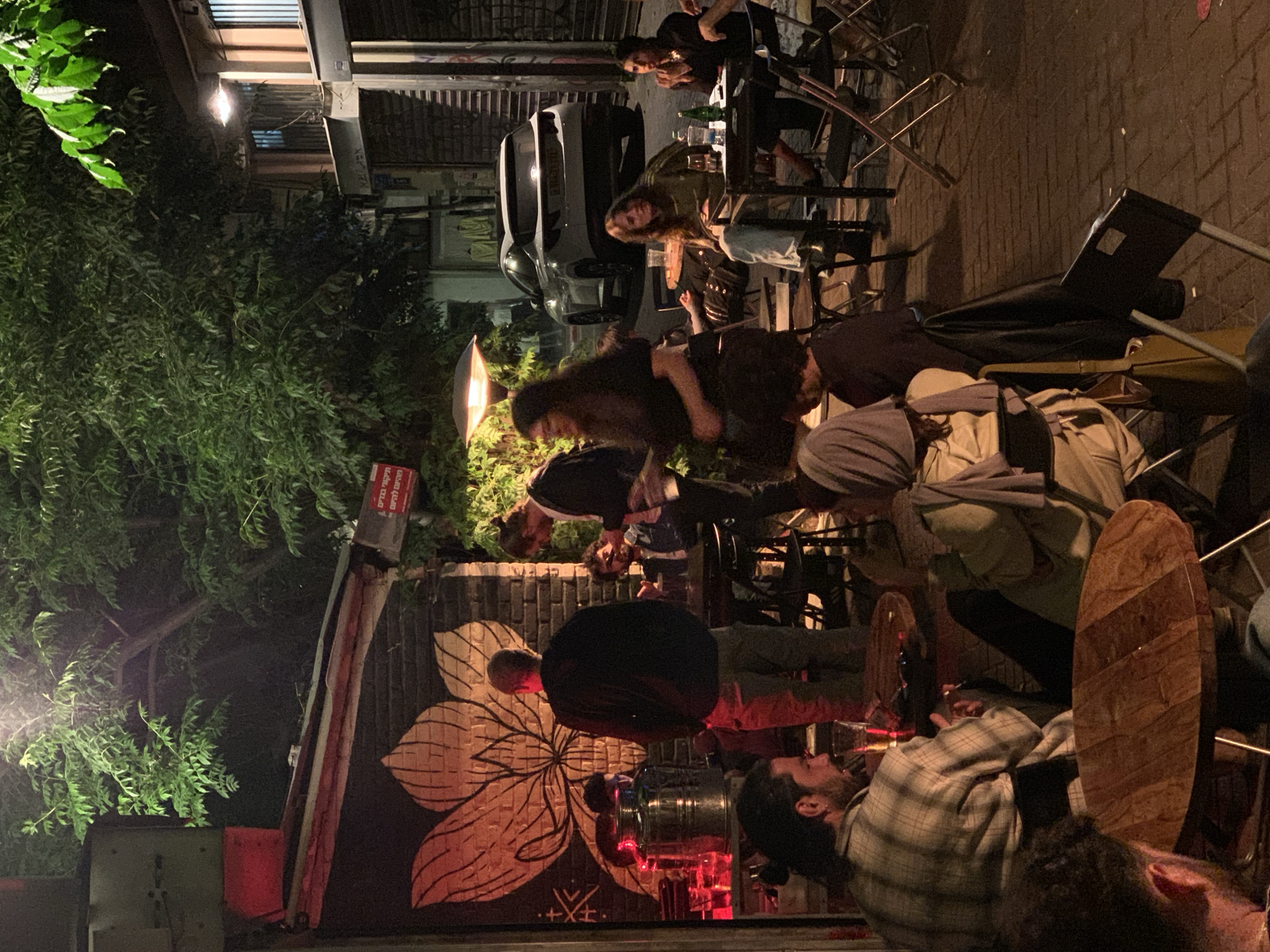The image captures a vibrant outdoor evening scene, likely a street-side bar or cafe in Tel-Aviv, Israel, filled with people socializing. The setting is illuminated by artificial lights, including street lamps, string lights, and overhead fixtures, indicating it is nighttime.
Numerous individuals, appearing to be young to middle-aged adults, are gathered in various groups. Many are seated at round tables with wooden tops and metal frames, while others stand or mill about. Drinks, including bottles and glasses, are visible on tables and at a bar counter. One person in a plaid shirt is seen in the lower right, looking upwards. A large, metallic beverage keg and red-lit glasses are noticeable at the bar area to the left.
The scene is backed by an urban environment, featuring buildings with shutters and a white car parked on the street. Lush green foliage from trees and bushes flanks the left side of the image, contrasting with the man-made structures. A significant feature is a large, stylized mural of an orange or red flower painted on a dark wall.
A prominent red sign with white Hebrew text is visible on the left, displaying phrases such as "מבצעים מיוחדים" (Special offers), "התקשר בכיף" (Call with pleasure), and "קמפיין" (Campaign). While other text might be present on distant shutters, it is largely illegible. The overall atmosphere appears casual and social, characteristic of an evening gathering in a lively urban spot.