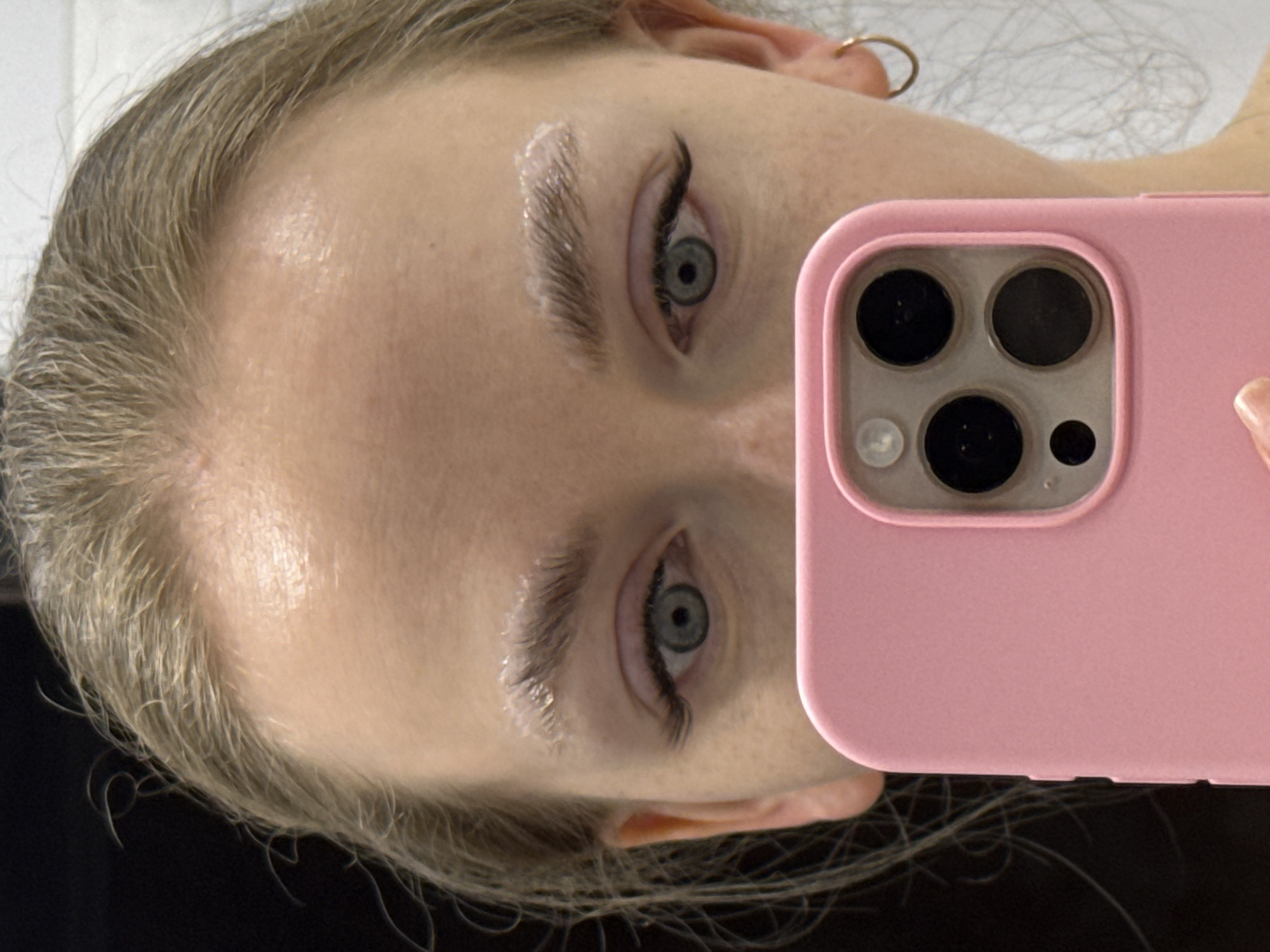 A close-up, indoor selfie presents a person with fair skin, light blonde hair, and blue-gray eyes. The person is holding a pink smartphone case close to their face, obscuring part of the view. The camera lens assembly on the phone is prominent. The person's eyebrows appear to have a clear gel or styling product applied. The lighting appears to be artificial. The location is not discernible. There is no indication the photo was taken in Petropavlovsk-Kamchatsky, Russia.