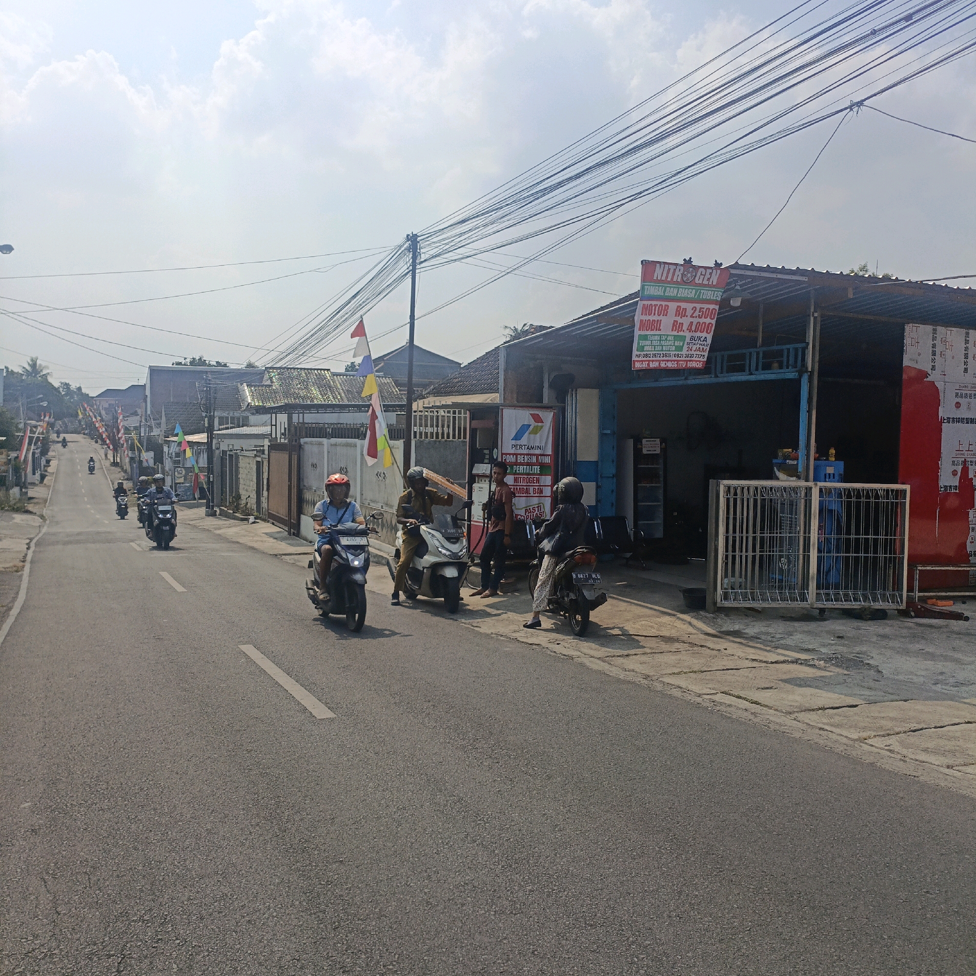 The image captures a street scene in Salatiga, Indonesia, on a bright, partly cloudy day. The road, asphalted and marked with a white line, stretches into the distance, flanked by buildings on either side. On the right, a workshop or service station is visible with a blue and metal facade, featuring a red sign that reads "NITROGEN" with additional text indicating services and prices for motorcycles and cars. A Pertamina gas pump is also present.
Several individuals on motorcycles are present. In the foreground right, a person wearing a blue shirt and a red helmet rides a dark-colored scooter. Further up the road on the left, another person in a blue shirt and a light-colored helmet is on a similar scooter, followed by two more motorcycles with riders. In front of the service station, one person stands near the gas pump, while another sits on a white scooter, and a third is on a dark scooter facing away from the camera.
The environment includes overhead electrical wires crisscrossing the sky, poles supporting them, and various flags, including Indonesian flags (red and white), displayed along the street and on buildings. The buildings appear to be of modest construction, typical of a residential or commercial area in Indonesia. Trees and vegetation are visible in the background, suggesting a suburban or town setting. The overall atmosphere is one of everyday activity on a street in Salatiga.