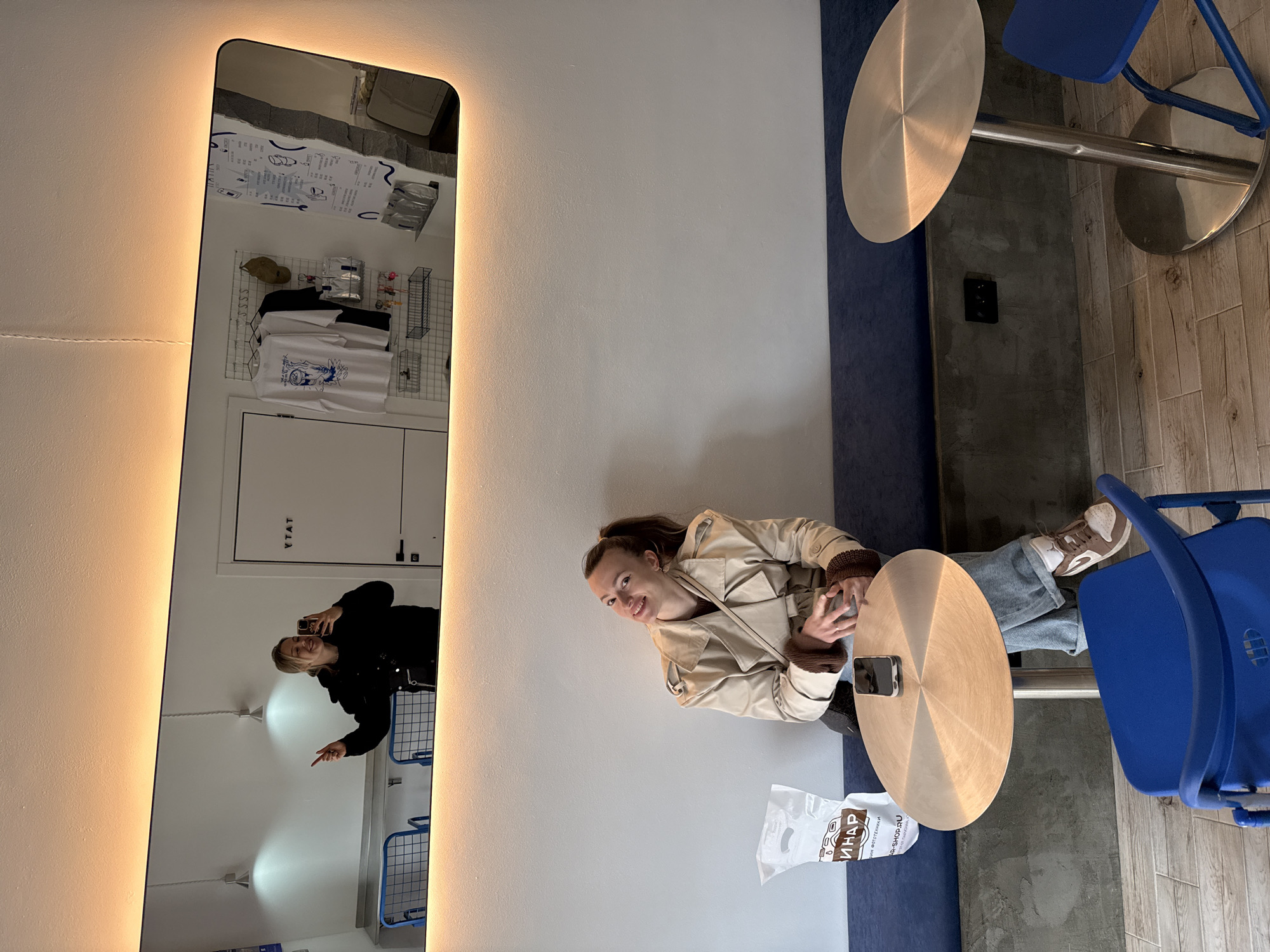 A young woman sits at a circular table in a restaurant, possibly in Krasnoyarsk, Russia. She is wearing a beige trench coat with brown knit detailing and light blue jeans. She is looking at the camera, smiling, with her hands near a smartphone on the table. A shopping bag with Cyrillic text is on the floor next to her. The table is next to a blue upholstered bench and a blue chair. In the background, a mirror reflects the interior of the restaurant, showing a person taking a photo, a white door with Cyrillic text, and various items for sale displayed on a wire rack. The lighting is bright, with a warm glow emanating from the mirror's frame.