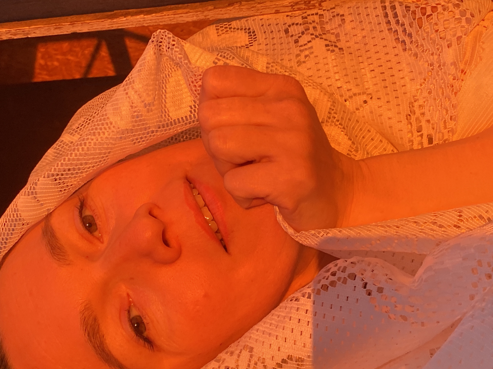 A close-up, high-angle portrait features a person, likely female, lying down and looking towards the camera with a gentle, slight smile. The scene is bathed in a warm, golden-orange light, indicative of either sunrise or sunset, which casts strong hues across the skin and surrounding elements.
The person's face is prominently displayed, framed by a white, loosely woven, mesh-like fabric, possibly lace or netting, which covers their head and parts of their body. Their eyes are open, revealing brown irises, and their mouth is slightly agape, showing teeth. A hand, clenched into a fist, rests near the left side of their face, partially covered by the fabric.
In the upper left background, a dark, wooden surface is visible, upon which angular shadows are cast, suggesting light filtering through a window frame or a similar structure. These strong shadows also fall across the textured white fabric, highlighting its intricate open-weave pattern. The warm lighting creates a serene and intimate atmosphere, emphasizing the interplay of light, shadow, and textural details. No text is visible in the image.