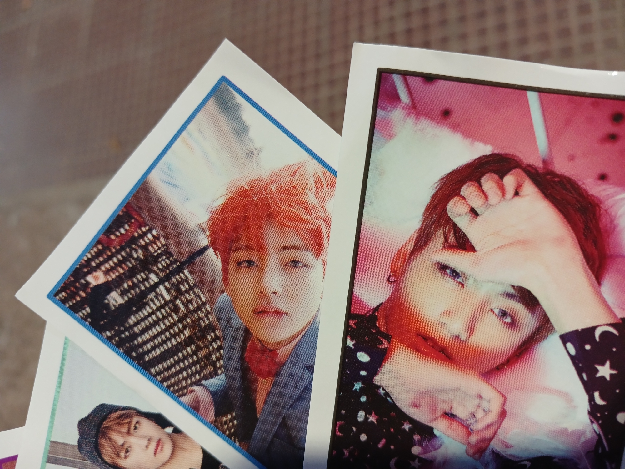The image shows three K-pop idol photo cards. The top left card features a person with orange hair and a blue suit with a red bow tie. The background appears to be a cityscape at night. The top right card depicts a person with dark hair, wearing a shirt patterned with stars and moons. The person is lying down with their hand near their face. The bottom left card shows a person with a beanie, partially visible. The background of the image is a blurred surface.