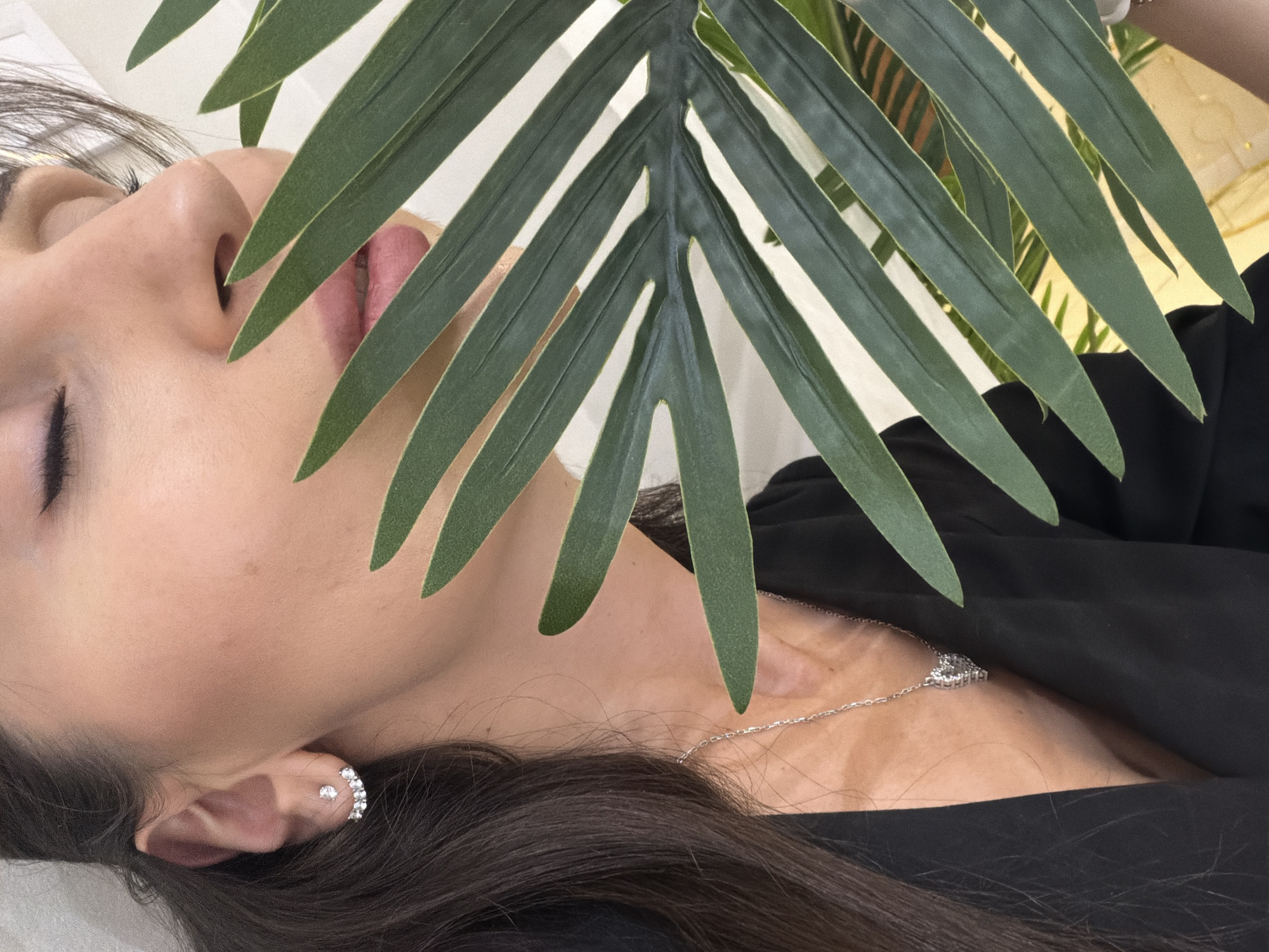 A close-up, angled portrait features a person, likely female, with her eyes gently closed, conveying a sense of serenity or repose. Her face, characterized by fair skin and light pink lips, is partially framed and softly shaded by a large, dark green, palm-like leaf positioned diagonally across the upper portion of the frame. Her dark hair cascades around her neck and shoulder.
She is dressed in a black garment and accessorized with silver-toned jewelry. A delicate necklace with a sparkling, possibly heart-shaped, pendant rests on her neck. Her visible ear is adorned with two distinct pieces: a small, round, clear gemstone stud and a curved ear cuff encrusted with multiple small, clear gemstones. The leaf itself is prominently veined, adding to the organic aesthetic of the shot.
The background is a soft, indistinct light color, appearing to be a wall or backdrop, with blurred hints of warmer, golden tones and possibly more greenery in the distance. The lighting is soft and diffused, creating a calm and intimate atmosphere. No text is visible within the image.