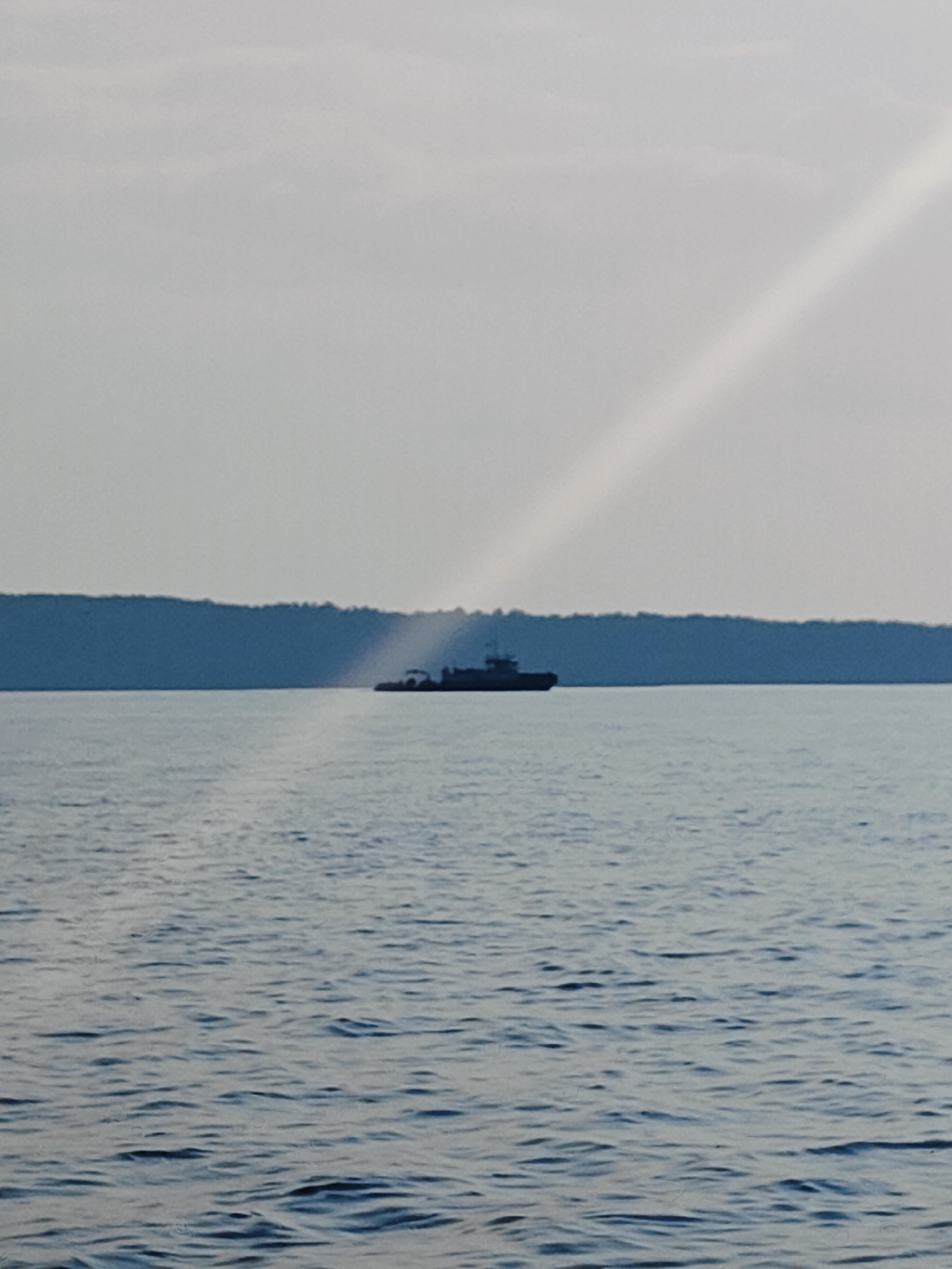 This media file captures a tranquil scene on a body of water, likely a lake or a wide river, under an overcast sky. The dominant colors are cool blues and grays, with a soft, diffused light.
The main subject is a large, dark boat silhouetted against a distant, dark blue shoreline. The boat appears to be a ferry or a barge, with visible structures on its deck, suggesting it might be carrying cargo or passengers. The shoreline is a long, unbroken line of what seems to be dense forest or treeline, receding into the distance.
A striking diagonal beam of light cuts across the upper right portion of the image, breaking through the clouds and illuminating a section of the sky. This light source suggests that the sun might be low in the sky, possibly indicating either early morning or late afternoon. The water itself is calm, with gentle ripples reflecting the muted light.
The overall mood is one of peacefulness and solitude. The vastness of the water and the distant, indistinct shoreline contribute to a sense of serenity. There are no visible people, and the only object of focus is the lone vessel. The location context of Chaykovsky, Russia, suggests this might be a scene on the Kama River or one of the reservoirs in the region. There is no visible text in the media file.