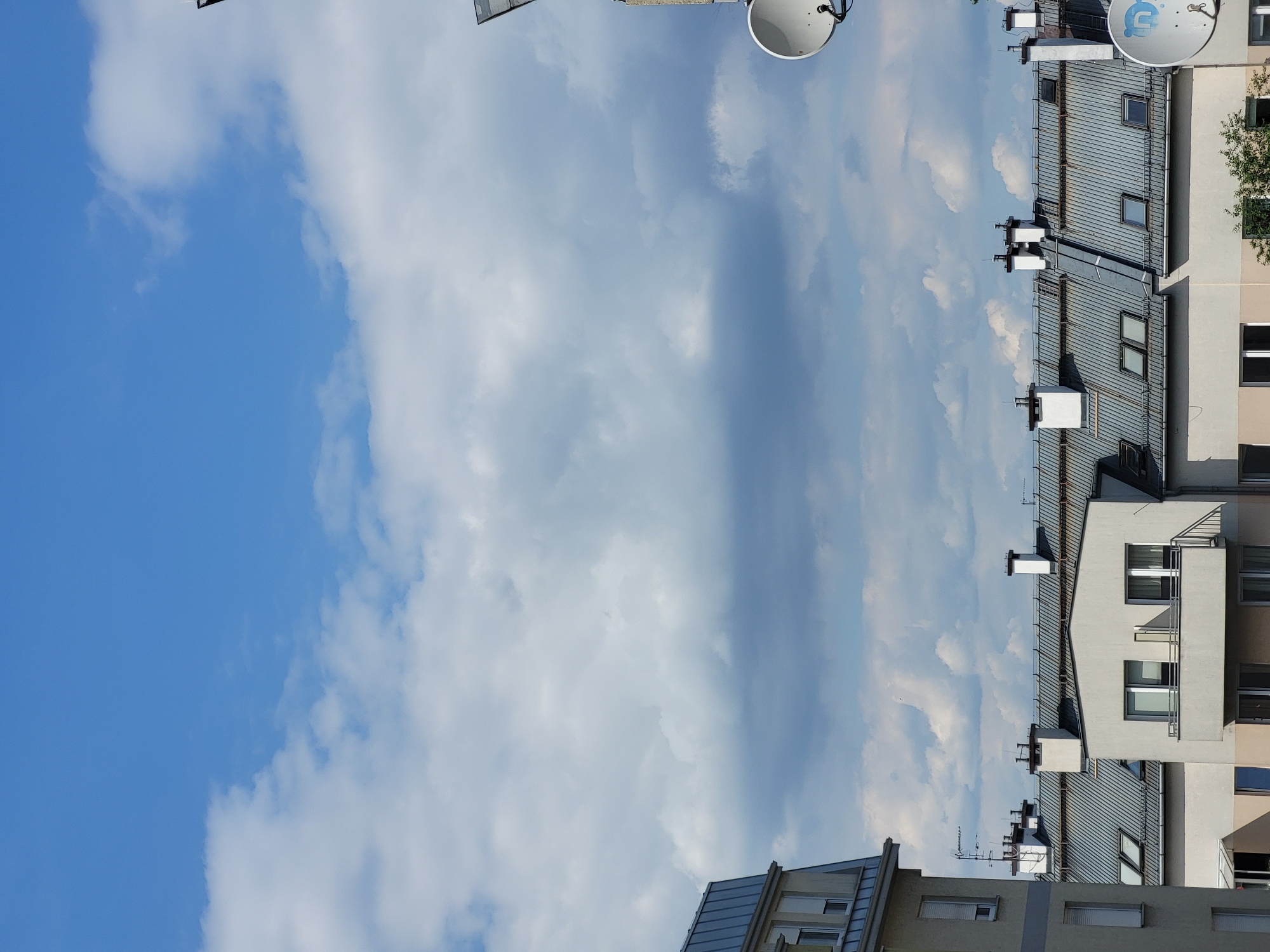 The image shows a view of rooftops and the sky in an urban environment. The rooftops are predominantly gray and metallic, with some featuring chimneys and skylights. Several satellite dishes are visible, attached to poles on some of the buildings. The sky is blue with scattered white clouds. The perspective suggests the photo was taken from an elevated position, looking down onto the buildings. The lighting indicates it is daytime, likely a bright, clear day with some cloud cover. There are no people visible in the frame. The scene appears to be a residential or commercial area.