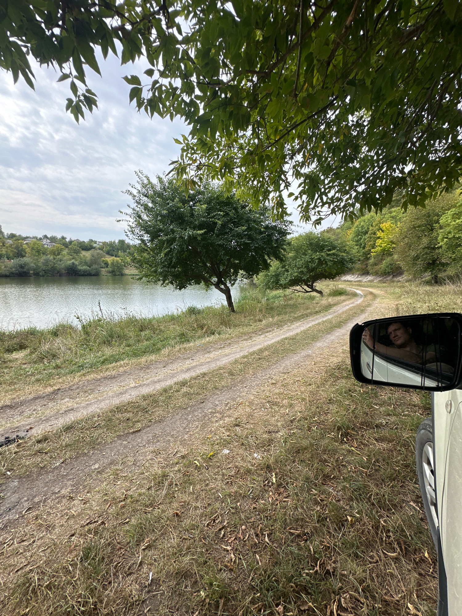 The image depicts a rural road alongside a body of water, likely a river or lake, with a person visible in the reflection of a car's side mirror. The road is unpaved, made of dirt and gravel, with tire tracks indicating recent vehicle passage. Dry grass and scattered fallen leaves line the roadside. Trees with green foliage dominate the landscape, with some exhibiting autumnal yellowing. In the distance, a gentle hillside is covered with more trees and a few scattered houses, suggesting a rural settlement. The sky is overcast with a soft, diffused light. The reflection in the car mirror shows a person's head and upper torso, appearing relaxed. The overall scene conveys a sense of natural beauty and tranquility. The presence of a road and houses suggests this location is likely on the outskirts of a village or town. Considering the provided context, this scene is located in Makiv, Ukraine.