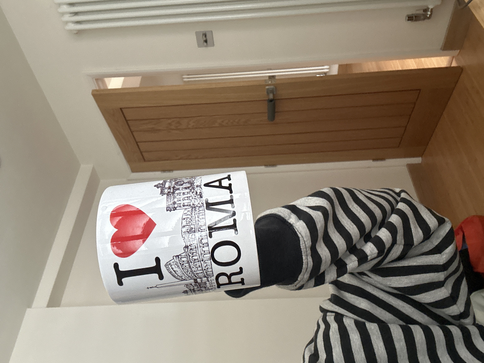 The image shows an "I <3 ROMA" mug held by a person wearing a black and white striped hoodie. The background includes a white wall, a radiator, a light switch, a wooden door with a metal latch, and a glimpse of a wooden floor. The location (Tunbridge Wells, UK) is not discernible from the image content.