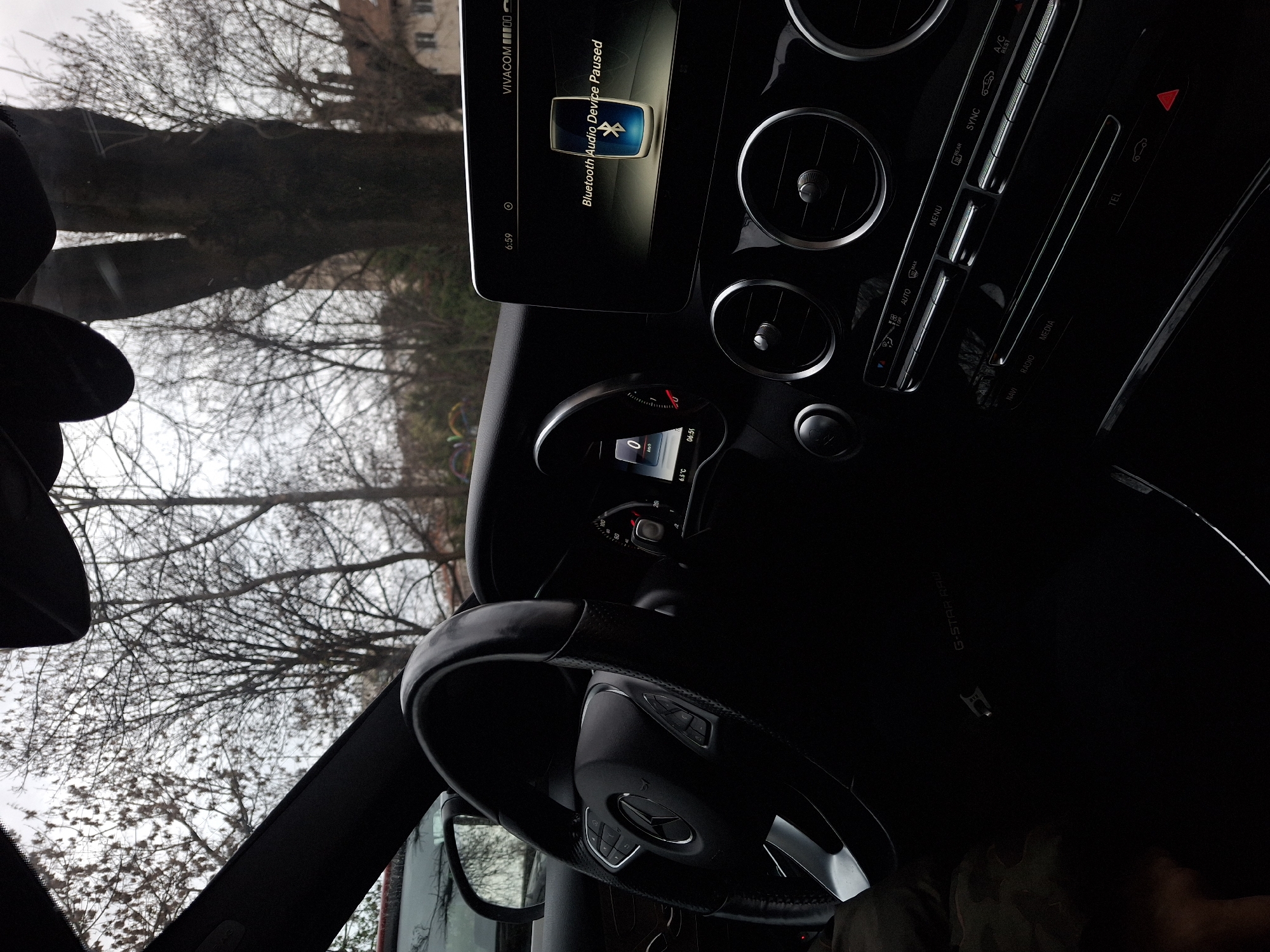 The image shows the interior of a car, specifically the dashboard, steering wheel, and infotainment system. The infotainment screen displays "Bluetooth Audio Device Paused" and indicates a time of 6:59. The car's climate control vents and buttons are visible to the right of the screen. The steering wheel has a Mercedes-Benz logo. Outside the windshield, trees with bare branches are visible. The display shows a temperature of 6.5 degrees Celsius and a time of 06:55. A "G-STAR RAW" logo is visible on the car's interior. The VIVACOM is noted on the display. The location is the city of Sofia, Bulgaria.
