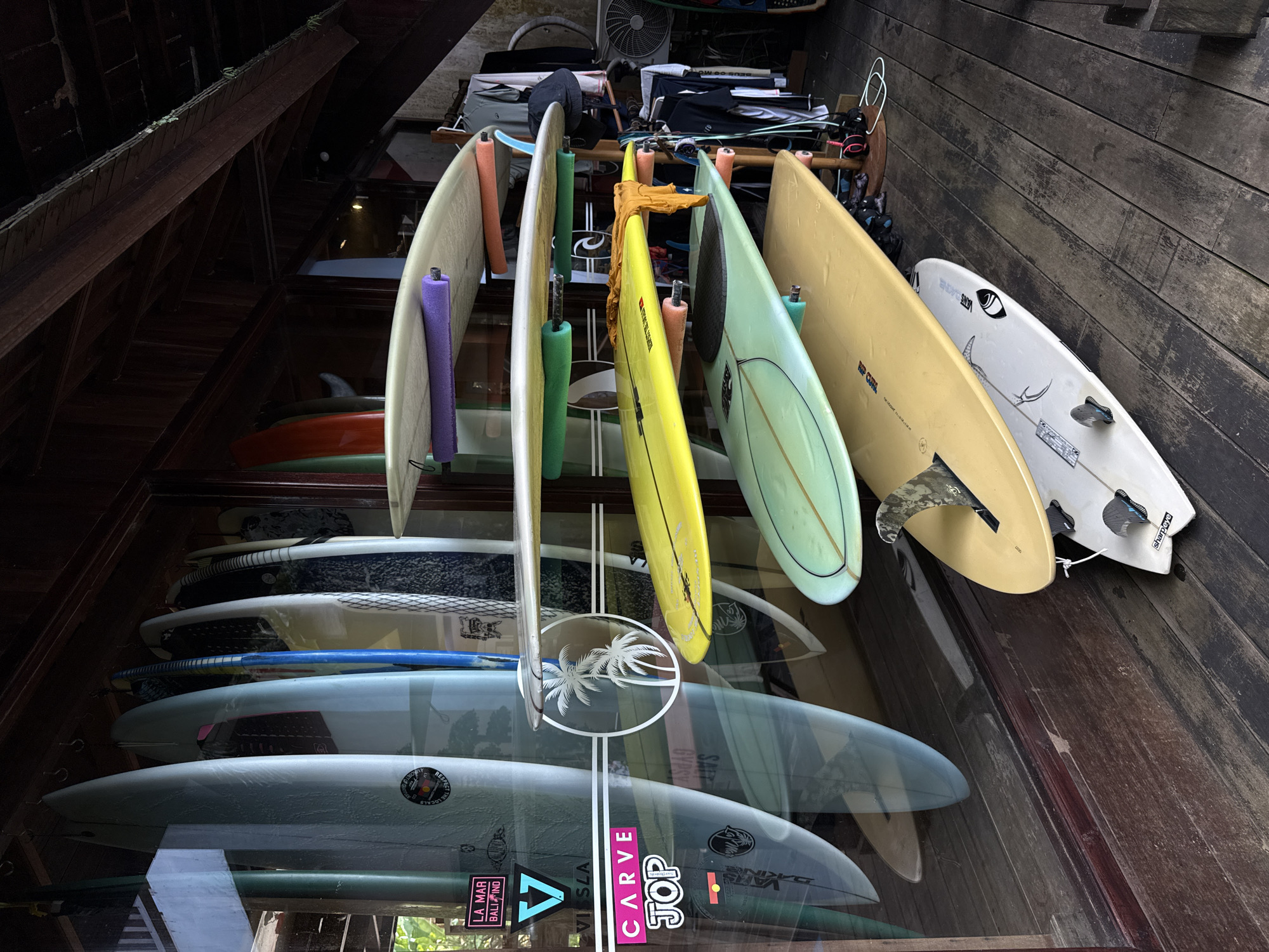 This image depicts a collection of surfboards stored in an organized manner. The surfboards are arranged in racks and stacked on shelves, with some boards positioned vertically and others horizontally. The surfboards exhibit a variety of colors and designs, with some featuring stickers or logos, including "RIP CURA," "GYPSY," "SAIT," "VISSLA," "CARVE," "JOP," "VANS DAGE," and "LA MAR BALI IND." The surroundings appear to be a storage area, possibly within a surf shop or a private collection. The scene is indoors, under a wooden roof structure, and the reflections on the surfboards suggest a glass enclosure. The location is possibly in a coastal town, but the city is Unknown, Unknown.