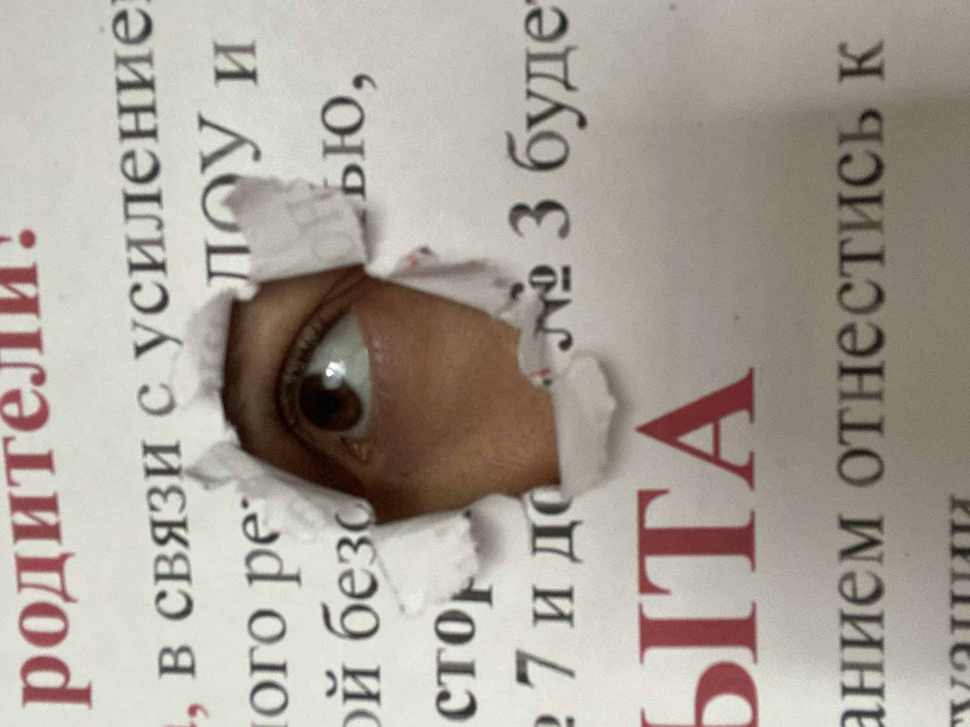 A human eye with a brown iris and a glint of light is seen peering through a ragged, irregularly torn hole in a piece of white paper. The torn edges of the paper are visible, framing the eye. The paper features printed text in black and some in red, suggesting it might be a document, newspaper, or book page. The text is oriented vertically, implying the paper was held or photographed at an angle.
Visible Russian text fragments include: "родители." (parents.), "1, в связи с усиление" (1, in connection with the strengthening), "ого ре", "ой безо", "Стор", "Лоу и", "Дью,", "27 и до № 3 буде" (27 and until No. 3 will be), "ЫТА", and "анием отнестись к" (handle with care / relate to). The act of looking through the torn paper suggests curiosity, observation, or perhaps a playful act of peeking. The location context is Chaikovsky, Russia. The time of day and weather are not discernible from the image.