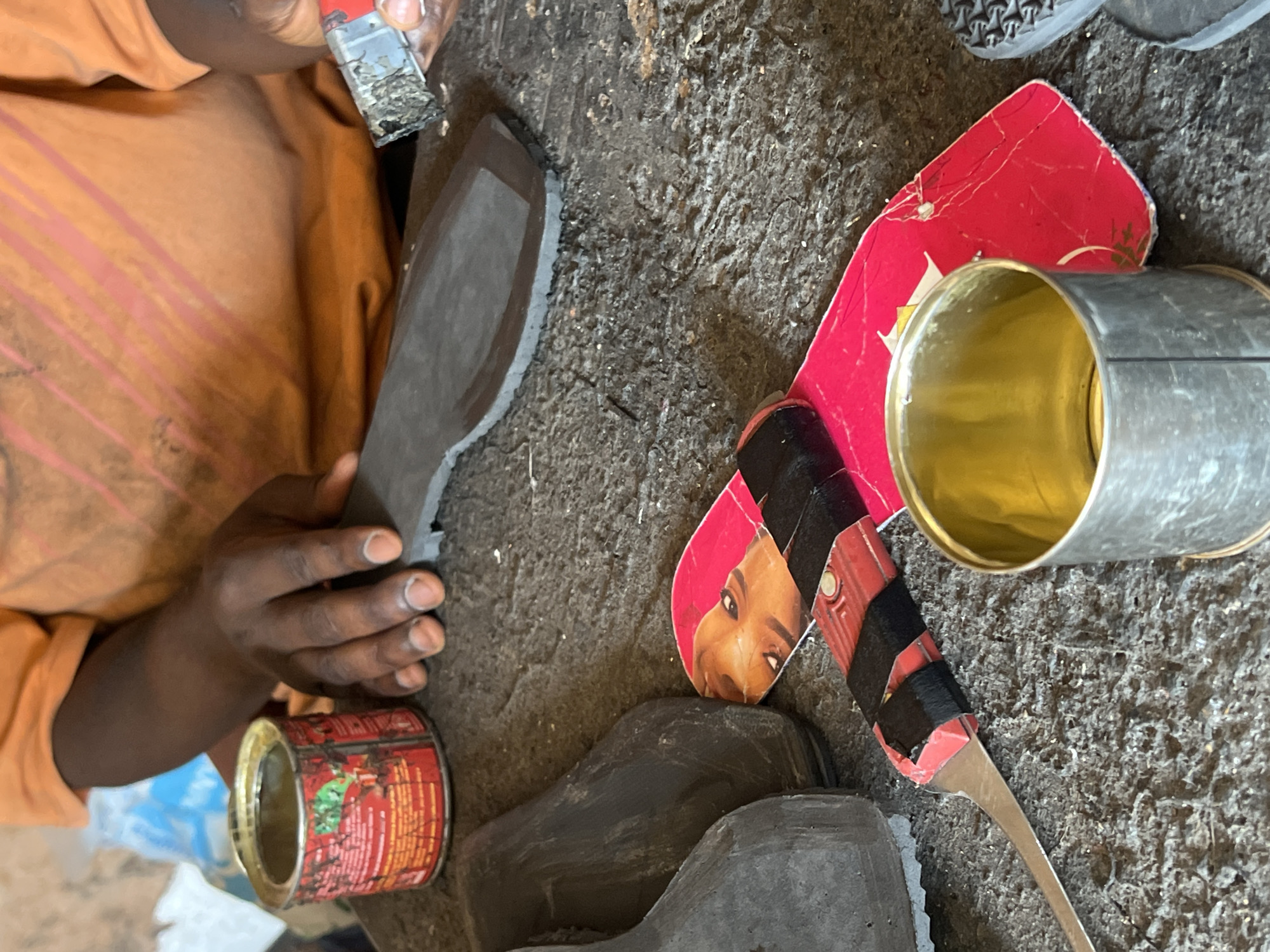 In a shoe repair workshop in Jos, Nigeria, a shoemaker works at a rough, stone-like workbench. The shoemaker, wearing an orange t-shirt, has dark skin and is shown with their hands, one of which is holding a small, metal tool with a blade covered in dark material. In front of them is a dark-colored shoe sole. To the left of the shoemaker are several dark shoe uppers, a red tin of what appears to be glue, and a piece of red cardboard with a picture of a person's face on it. In the foreground, a metal can, likely containing glue or polish, is positioned next to the red cardboard. The overall scene depicts a craftsman engaged in the detailed work of shoe repair.