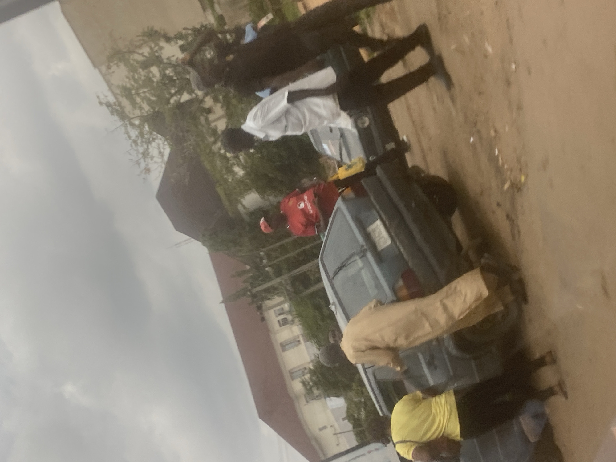 The image shows a group of people gathered around a light blue vehicle in Abuja, Nigeria. There are at least seven people visible. One person is wearing a red shirt and red cap. Another person is wearing a white shirt. A third person is wearing a yellow shirt. Others are wearing dark-colored clothing. The vehicle is parked on a dirt or gravel surface. Buildings are visible in the background. The sky is overcast.