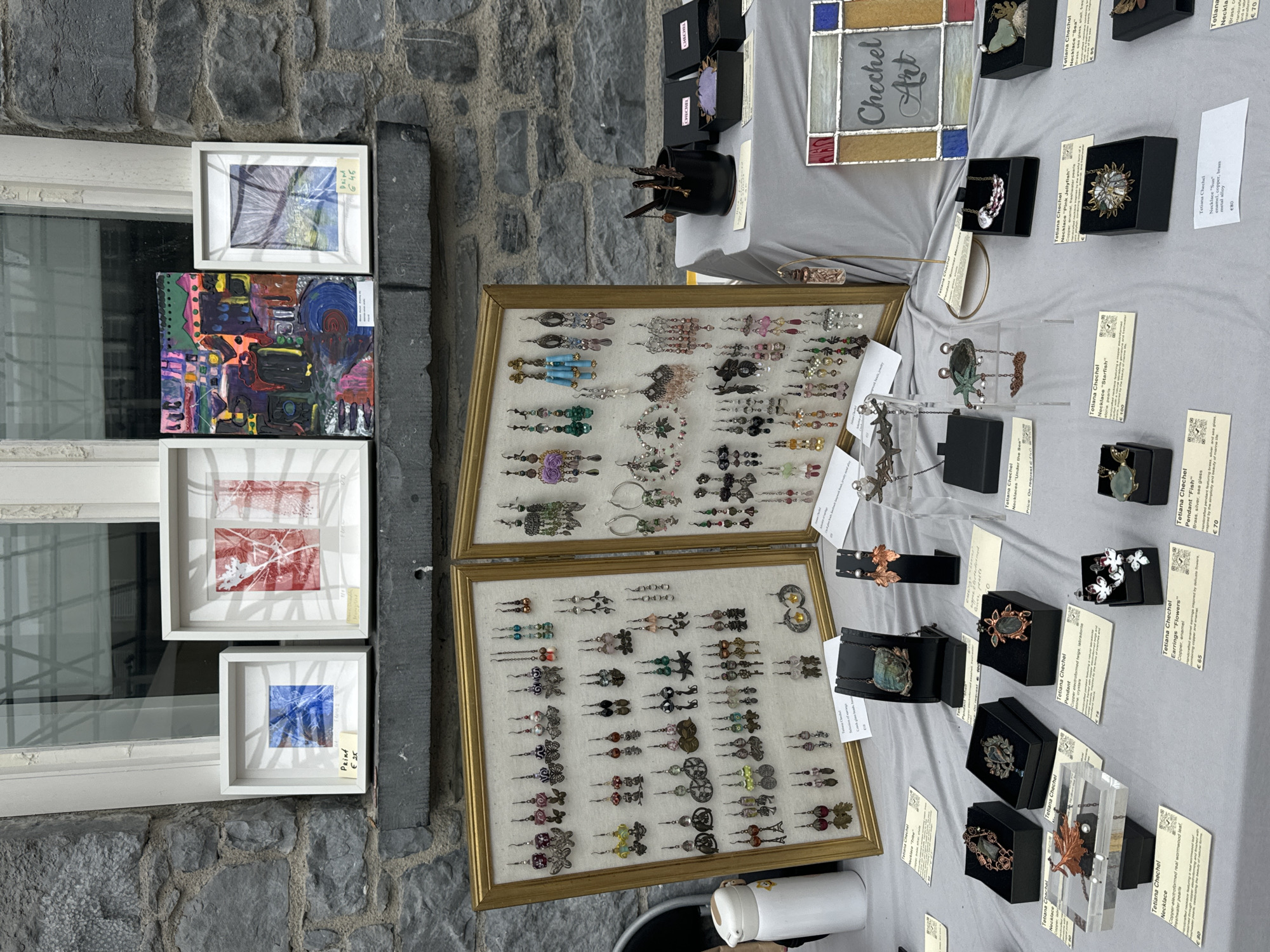 The image shows an outdoor market stall displaying handcrafted art and jewelry. The stall is set up against a stone wall with a window above. The merchandise includes framed prints and paintings on a shelf, and a large collection of earrings displayed on two framed boards. Below these, on a table covered with a grey cloth, are various pieces of jewelry, including necklaces and pendants, each with a descriptive tag. The overall scene suggests an arts and crafts fair or market. The signage "Chechel Art" indicates the name of the artist or vendor. While the location is mentioned as Kilkenny, Ireland, there are no specific landmarks visible in the image that would definitively confirm this location. There are no people clearly visible in the image.