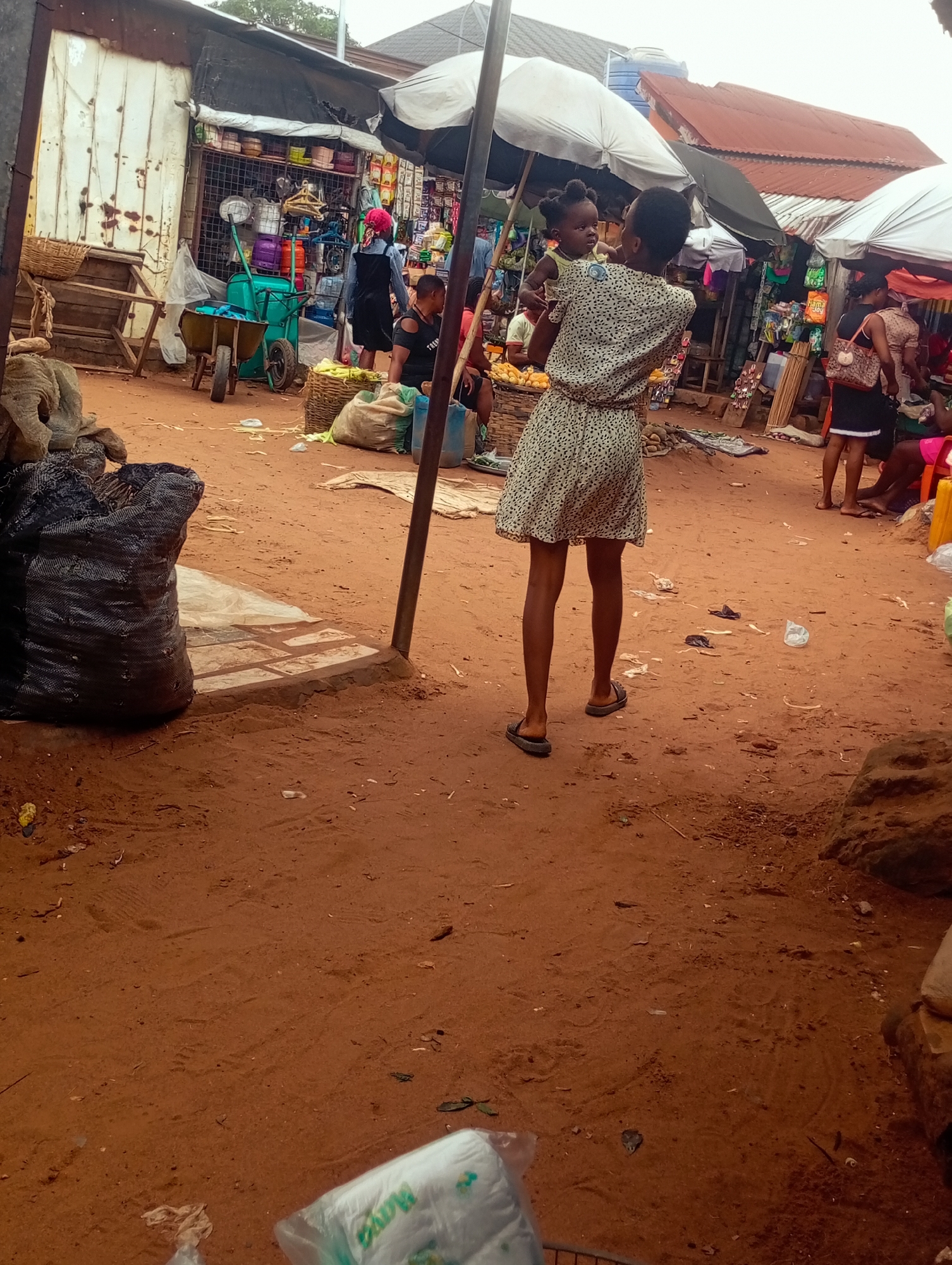 An outdoor market scene shows a woman holding a baby while standing in a dusty, reddish-brown dirt area. The woman is wearing a light-colored dress with a dark speckled pattern. The baby, facing away from the camera, is being held in the woman's arms. In the background, there are stalls with various goods displayed under awnings and umbrellas. Some of the stalls appear to be selling clothing, household items, and produce. A cart with wheels is visible near one of the stalls. Several other people are present in the market, though their features are not distinct. Large black sacks are placed on the ground in the foreground, and there is some scattered debris. The overall impression is that of a busy, informal marketplace.