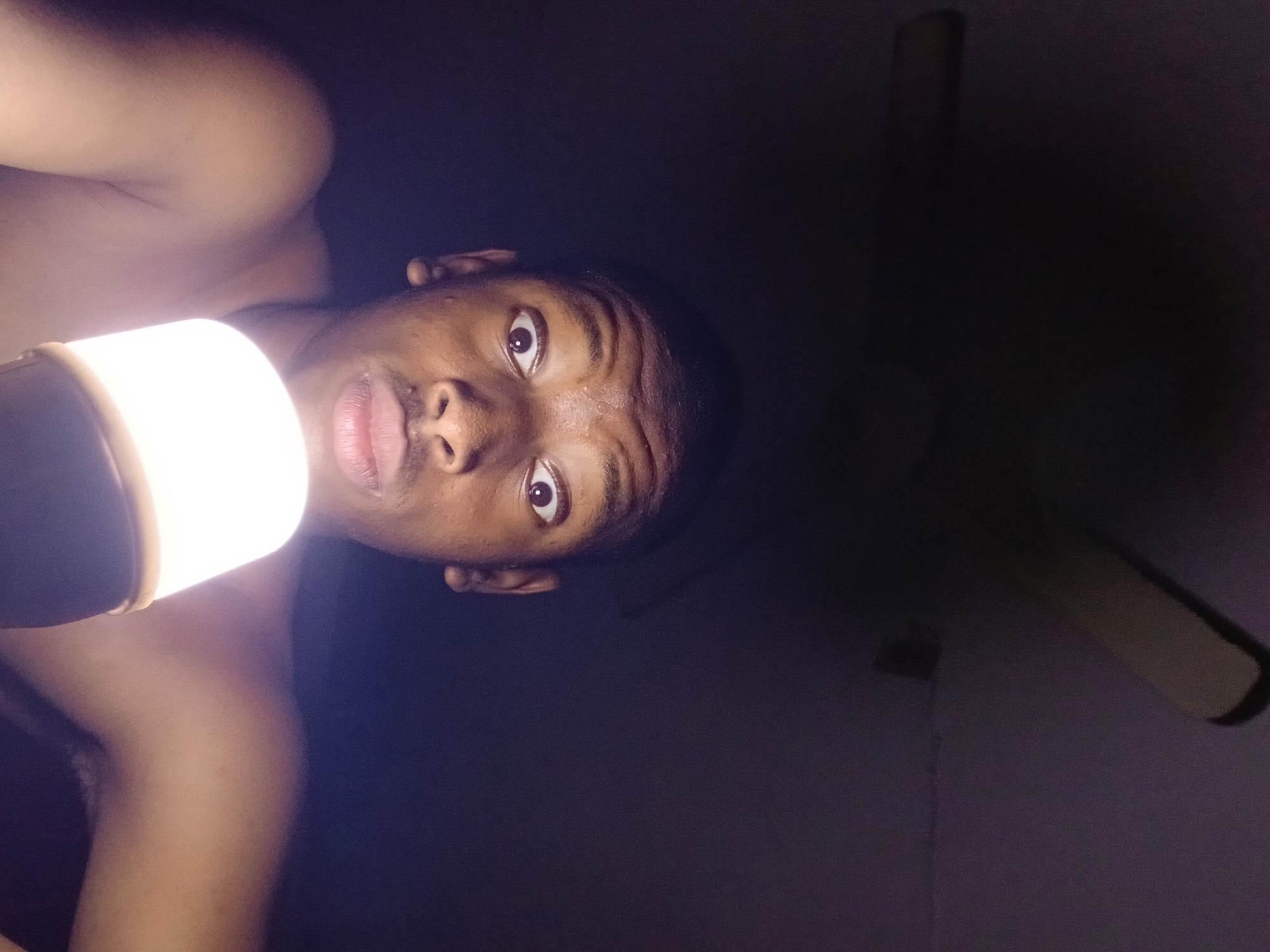 A young Black male is prominently featured in a dimly lit indoor setting, illuminated primarily by a bright, cylindrical light source held close to his face. He looks directly at the camera with wide, attentive eyes and a slightly open mouth, exhibiting a look of surprise or curiosity. Beads of sweat are visible on his forehead and nose. He appears to be shirtless or wearing light-colored attire that blends with the intense light.
The strong, direct artificial light creates harsh highlights on his face and upper body, casting deep shadows around him and contributing to the overall dramatic and stark quality of the image. The background is almost entirely dark, but the faint outline of a ceiling fan is discernible in the upper right, confirming an indoor location. The extreme darkness of the room, requiring a portable light source, suggests it is night or that there is a power outage. The activity taking place is likely a selfie or portrait taken in these low-light conditions. No visible text is present. The scene is set in Lagos, Nigeria.