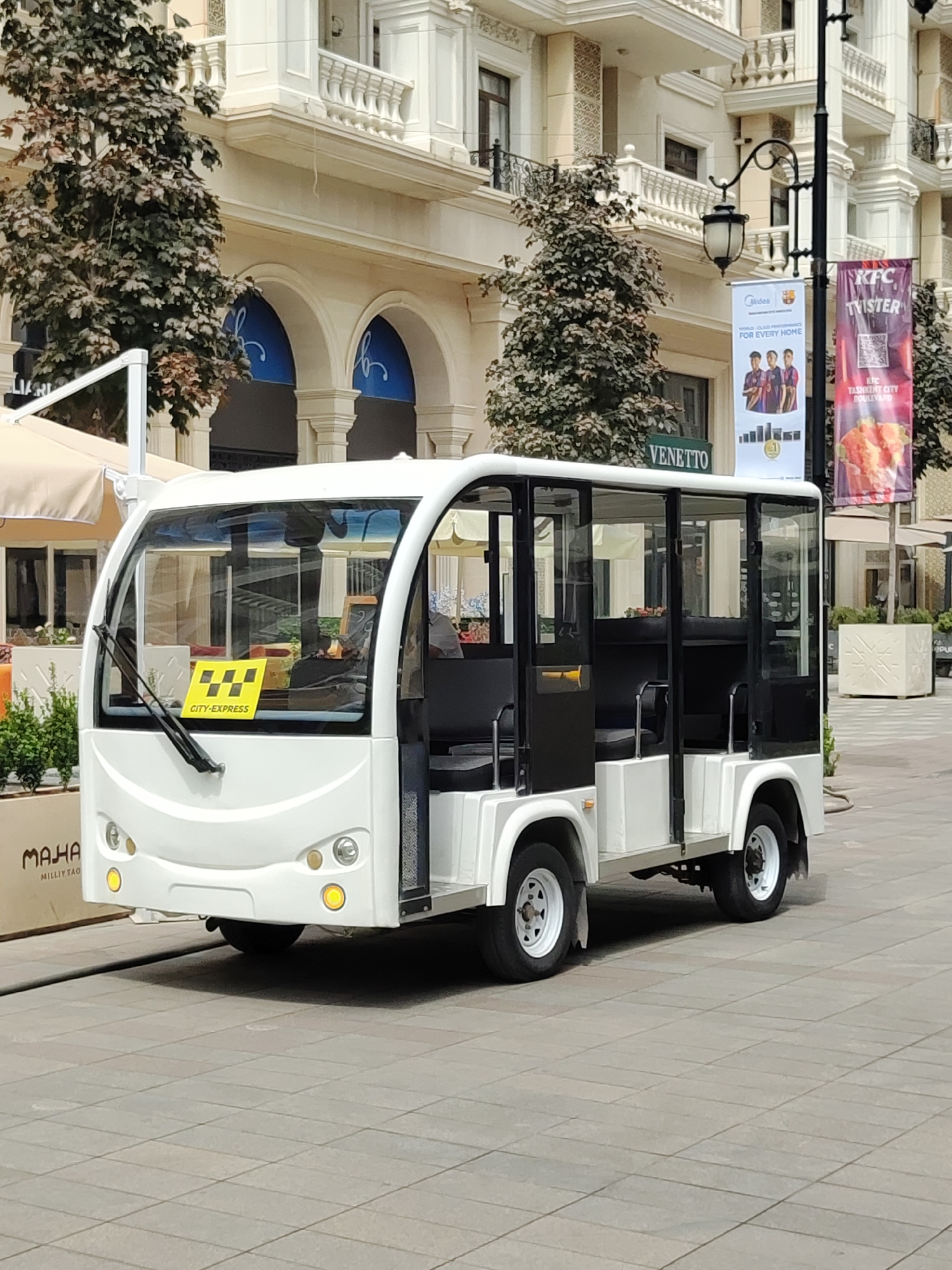 A white, open-air shuttle bus with black accents and seating is parked on a paved pedestrian walkway in front of a multi-story building with light-colored facade and arched entrances. The shuttle has a yellow sign on the windshield that reads "CITY-EXPRESS" with a checkered taxi symbol. Several trees with dark foliage are visible around the building. Banners hang from a lamppost on the right side of the image, one advertising "KFC TWISTER" and another with the "Midea" logo. The building and the overall setting suggest a commercial or urban area. The image was taken in Tashkent, Uzbekistan.