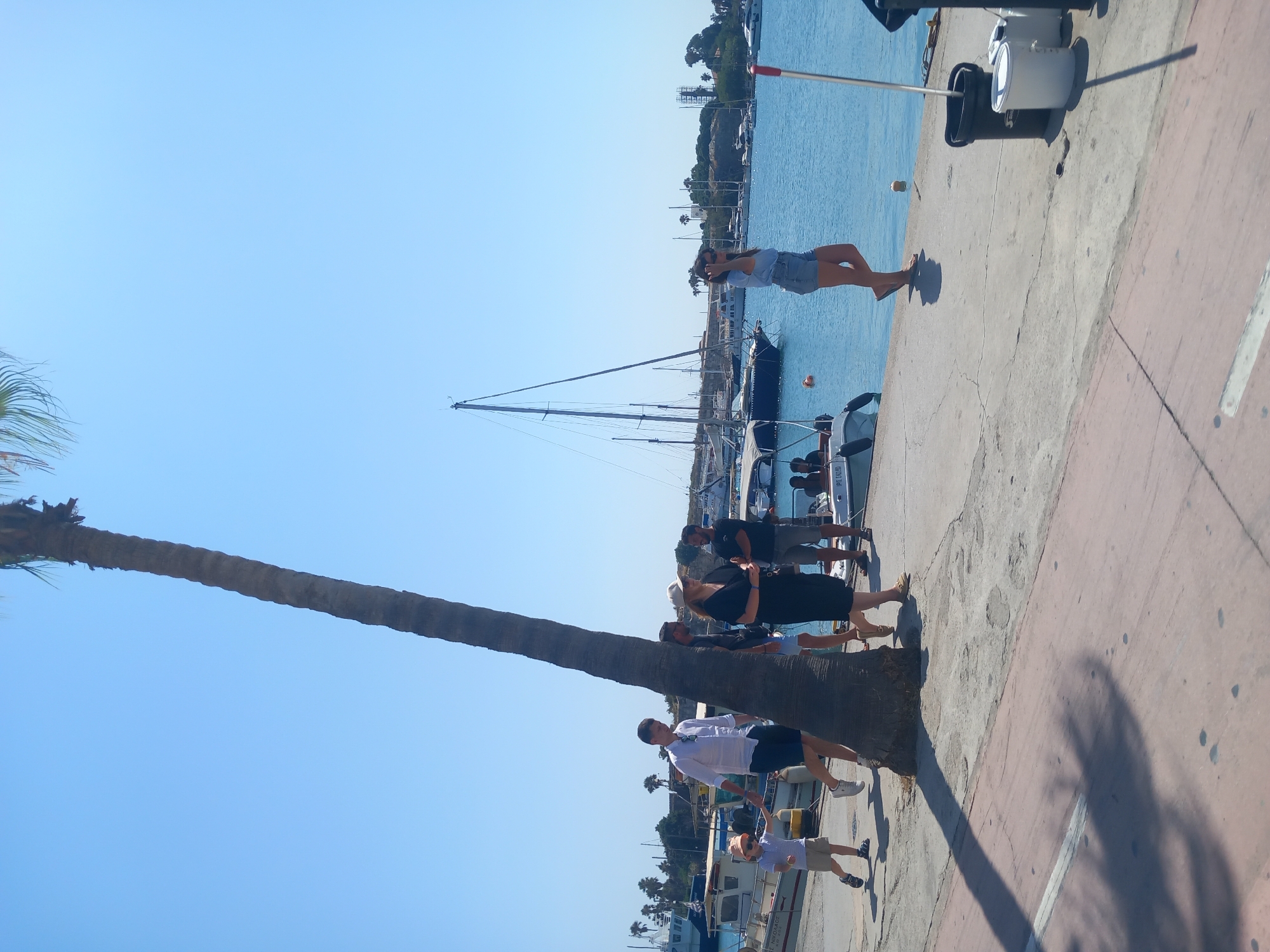The image depicts a scene at the harbor in Kos, Greece, under a clear blue sky. A tall palm tree dominates the foreground, casting a shadow on the paved walkway. Several people are visible walking along the harbor. On the left, a man in a white shirt and blue shorts walks alongside a young child wearing a striped shirt and shorts, and a cap. Further to the right, a group of individuals are walking, including a woman in a dark dress and a hat, and a man in a dark t-shirt and shorts. A woman in denim shorts and a light blue top is walking on the right side of the frame, looking away from the camera. In the background, various boats and yachts are docked in the blue water. The environment suggests a sunny day at a bustling port.