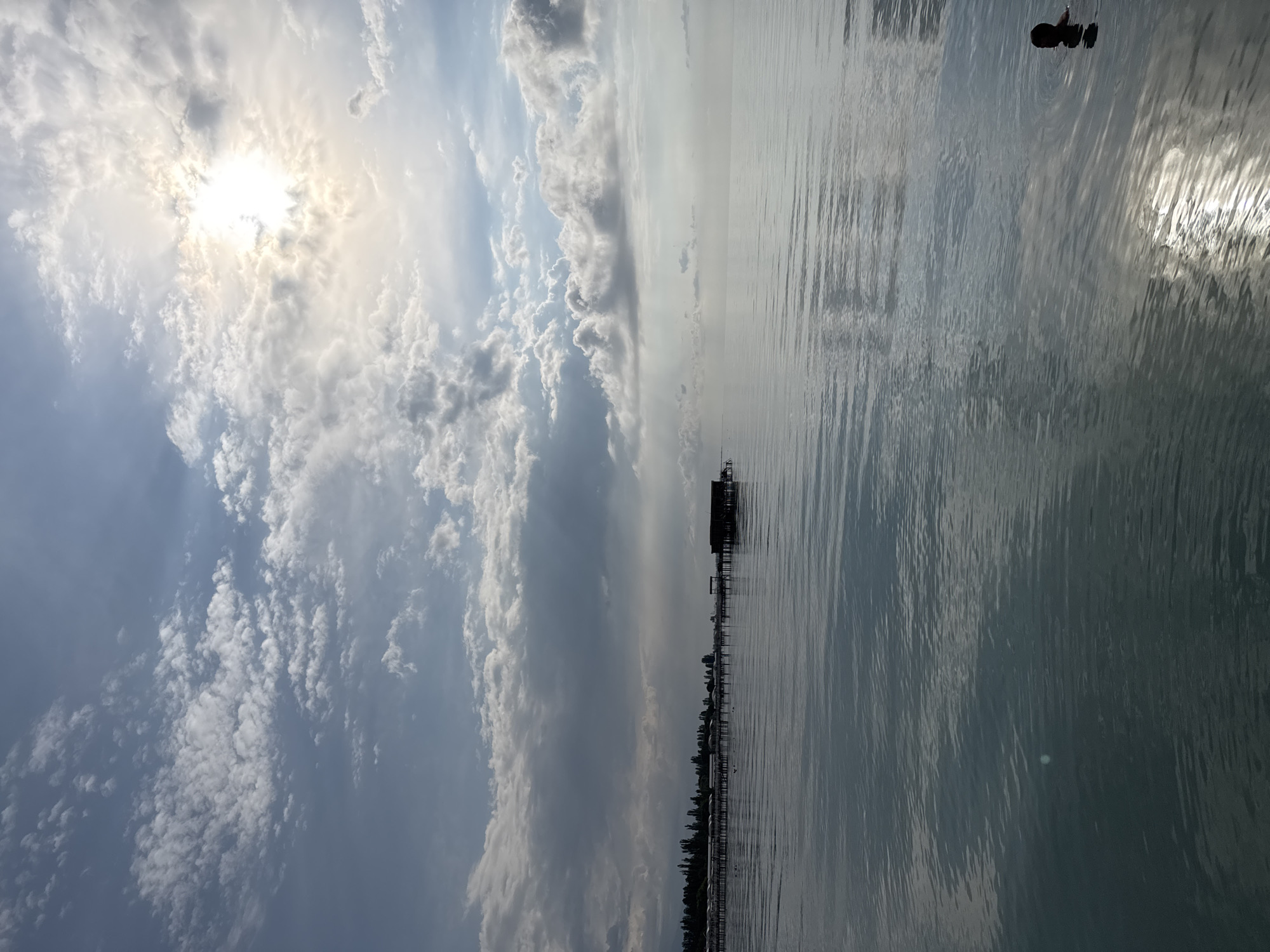 In the city of Sary-Oy, Kyrgyzstan, this image captures a serene view of the Issyk-Kul lake under a cloudy sky. The sun is partially obscured by clouds, casting a bright glow and reflecting on the calm water. In the distance, a long wooden pier stretches across the lake, leading to a small, dark structure, possibly a boathouse or fishing hut. The pier is lined with a series of smaller structures with rounded roofs, suggesting a resort or residential area. The shoreline is marked by a line of trees, creating a natural boundary between the land and the lake. In the foreground, a person is swimming in the lake, their head and shoulders visible above the water. The water is calm, with gentle ripples reflecting the sky and sunlight. The overall scene evokes a sense of peace and tranquility, with the vastness of the lake and the expansive sky creating a sense of natural beauty.