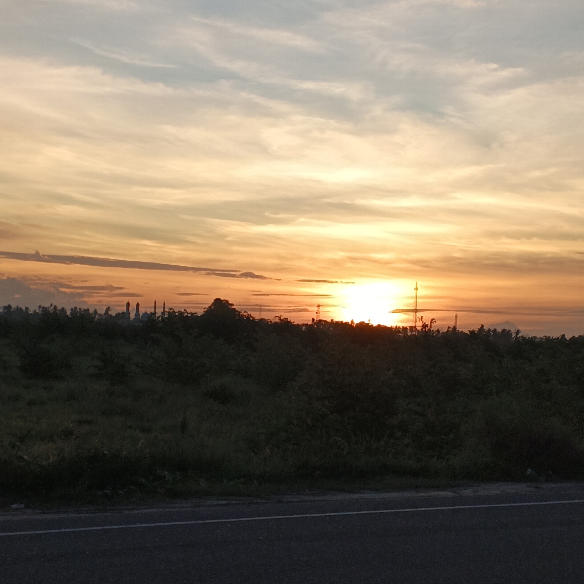 The image depicts a sunset in Dayah Kruet, Indonesia. The sky is painted with hues of orange, yellow, and pale blue, with wispy clouds stretching across the horizon. In the distance, silhouetted against the vibrant sky, are several prominent structures, possibly buildings or industrial stacks, and what appear to be domes, suggesting a town or religious center. Below the sky, a dark, dense line of vegetation forms the middle ground, with trees and foliage obscuring the ground below. In the foreground, a paved road with a white line marking separates the viewer from the natural landscape. The overall scene is tranquil, capturing the natural beauty of the sunset over the Indonesian landscape.