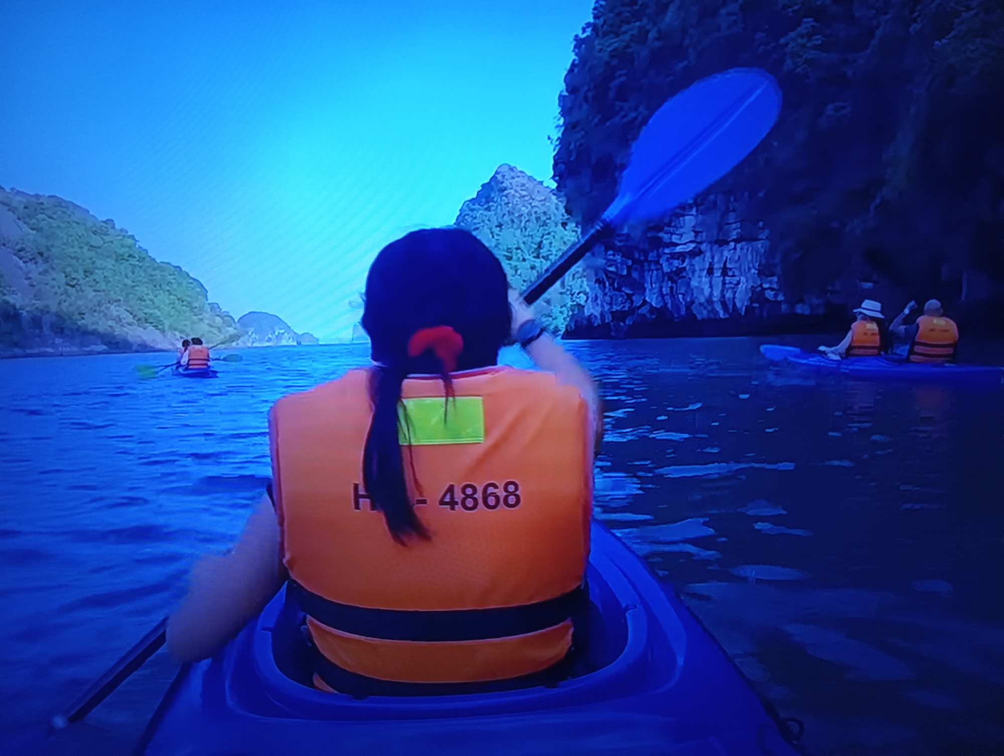 The image shows a person kayaking on a body of water, with limestone cliffs and lush vegetation on either side. The person is wearing an orange life jacket with the number "H - 4868" visible on the back. A blue kayak paddle is held by the person. In the background, other kayaks with people can be seen. The sky is clear and blue. The location is not Dnipro, Ukraine; this appears to be a scene from a tropical or sub-tropical environment, possibly Ha Long Bay in Vietnam, given the characteristic karst topography.