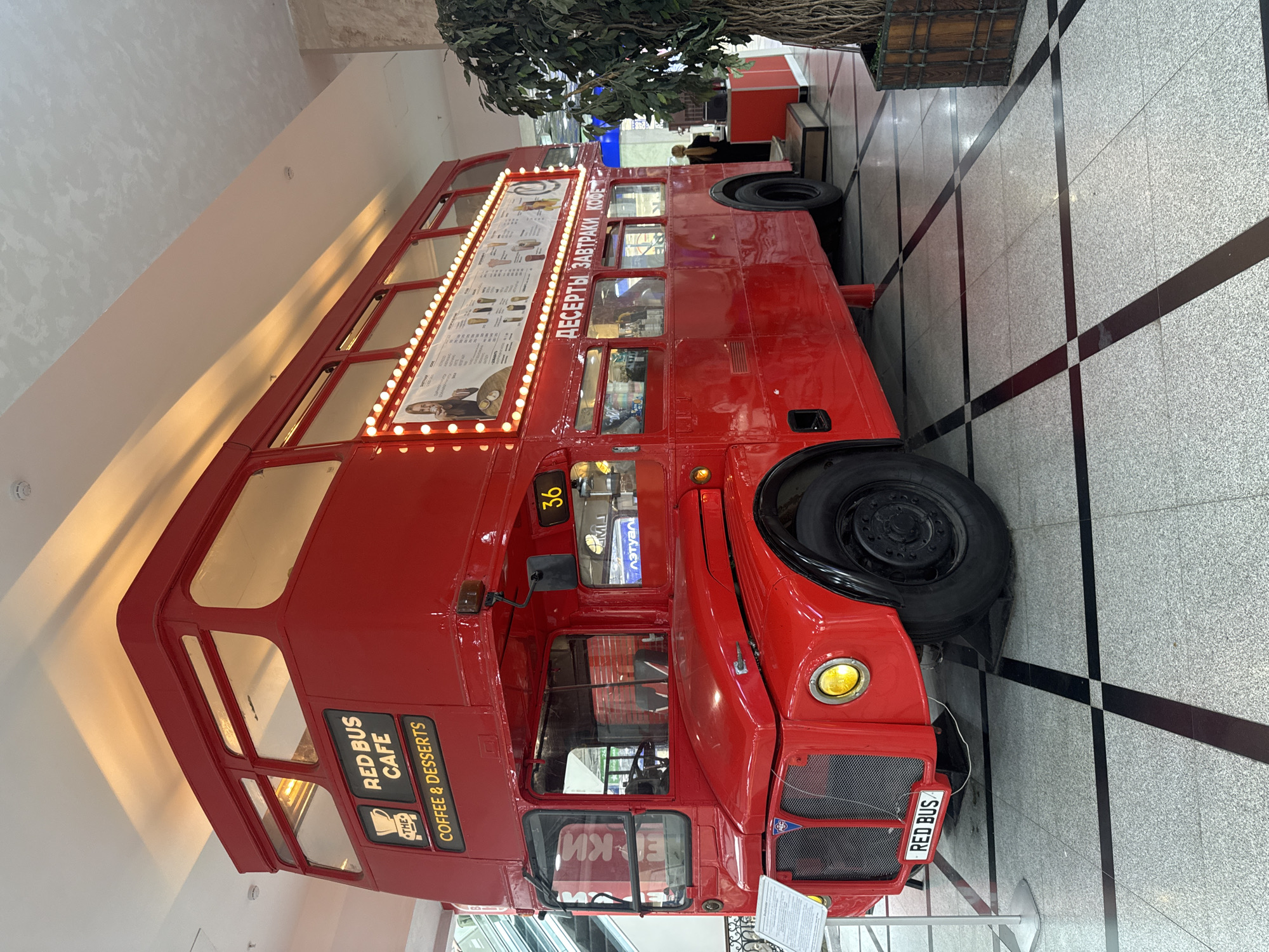 The image shows a bright red double-decker bus, modified to serve as a cafe. The bus is parked indoors on a patterned tiled floor, with black and grey tiles forming a checkered pattern. A large tree with artificial foliage is visible behind the bus, suggesting an indoor setting, possibly a shopping mall or entertainment venue.
The bus is adorned with numerous small light bulbs along the side of the upper deck, highlighting a menu board. The menu displays offerings in what appears to be Cyrillic script, with text such as "ДЕСЕРТЫ ЗАВТРАКИ КОФЕ" (Desserts Breakfast Coffee). There are also some images on the menu board, including what looks like a portrait of a person and various food or drink items.
On the lower front side of the bus, there are signs that read "RED BUS CAFE" and "COFFEE & DESSERTS," along with a coffee cup logo. A license plate on the front of the bus reads "RED BUS." The bus itself is a classic double-decker design, with large windows on both levels. The overall impression is that of a novelty cafe designed to attract customers with its unique theme. A person, partially visible in the background, appears to be standing near the bus. The lighting suggests it's daytime, as there are no artificial light sources clearly illuminating the scene from above, but rather ambient light creating reflections. The setting appears to be Yekaterinburg, Russia, as indicated by the Russian text on the menu board.